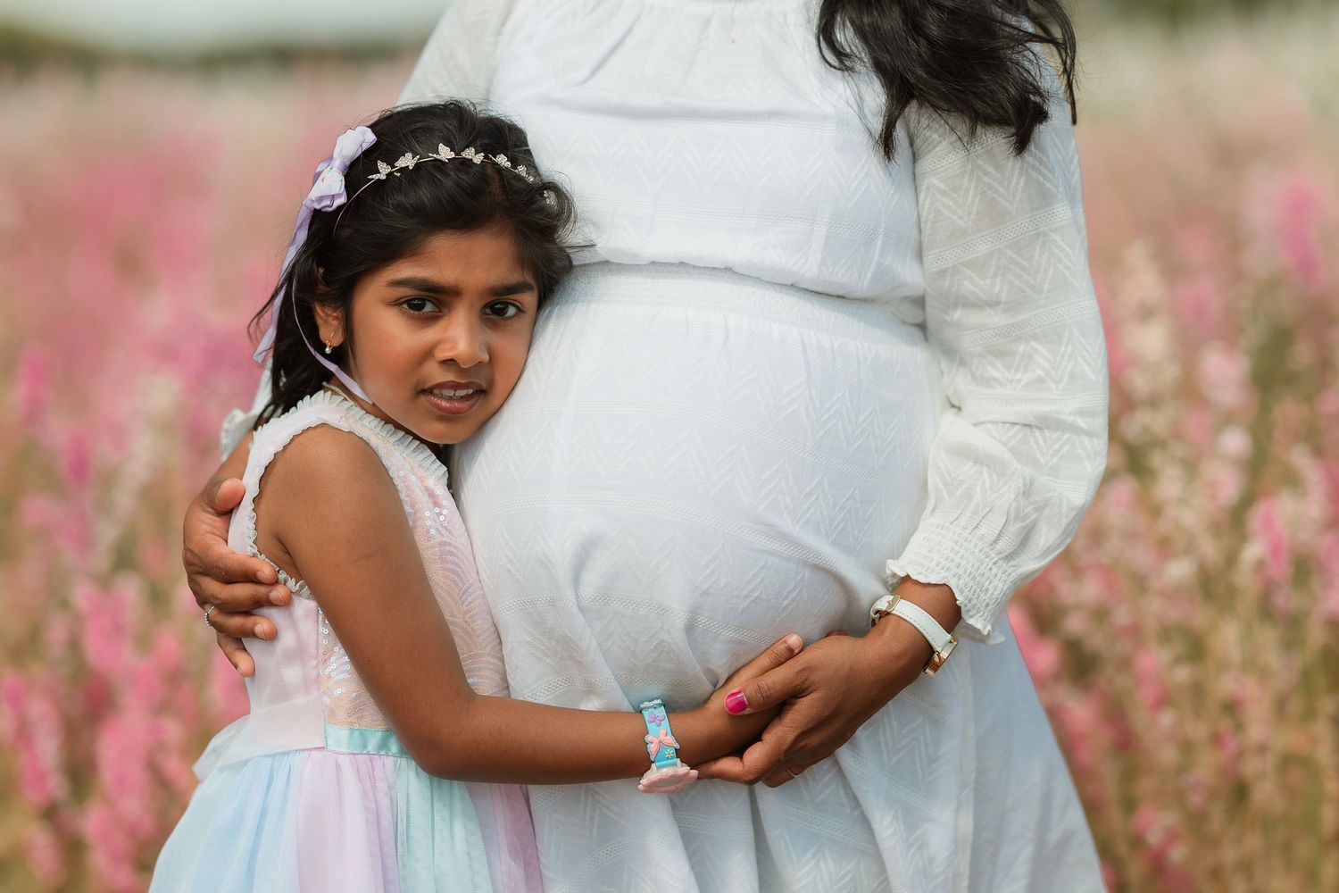 A maternity photo session in a field of pink wildflowers with someone wearing a white flowing dress.