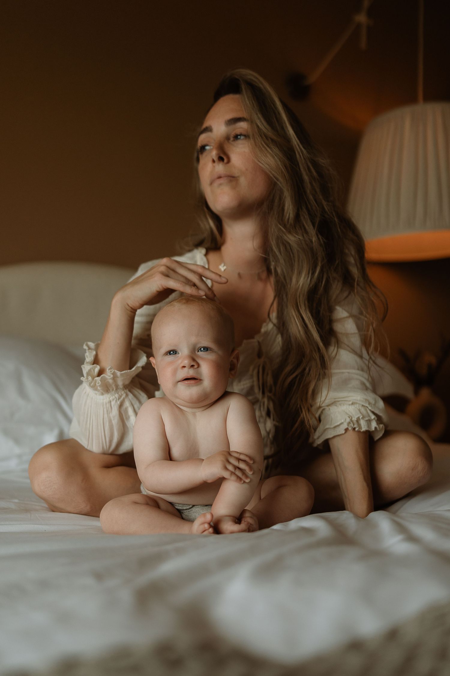 A tender moment captured between parent and baby lying together on white bedding in warm, intimate lighting.