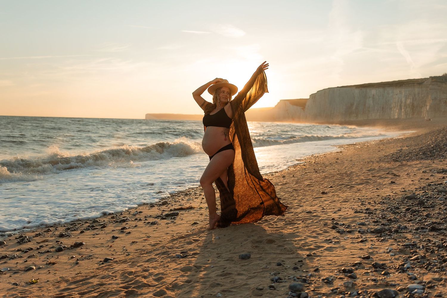 A person in a flowy dress poses gracefully on a sandy beach at sunset with ocean waves in the background.