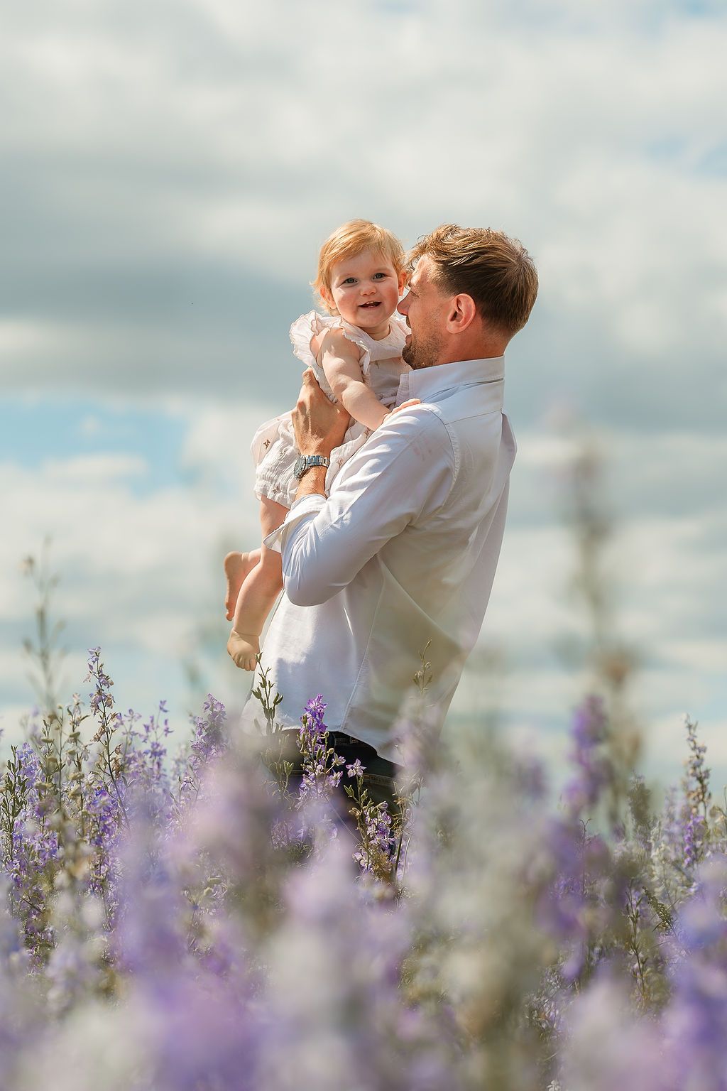 Father holds smiling baby girl in lavender field under cloudy sky, sharing a tender moment outdoors.