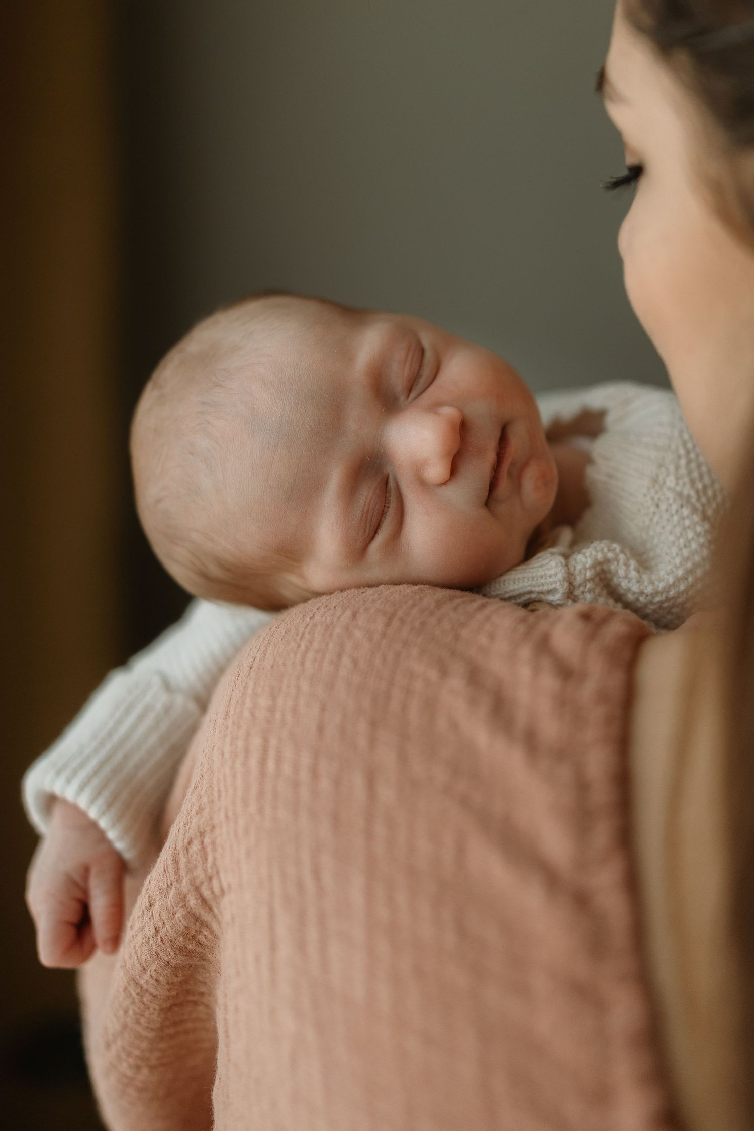 A peaceful sleeping newborn baby wearing a white knit sweater rests against someone's shoulder in soft lighting.