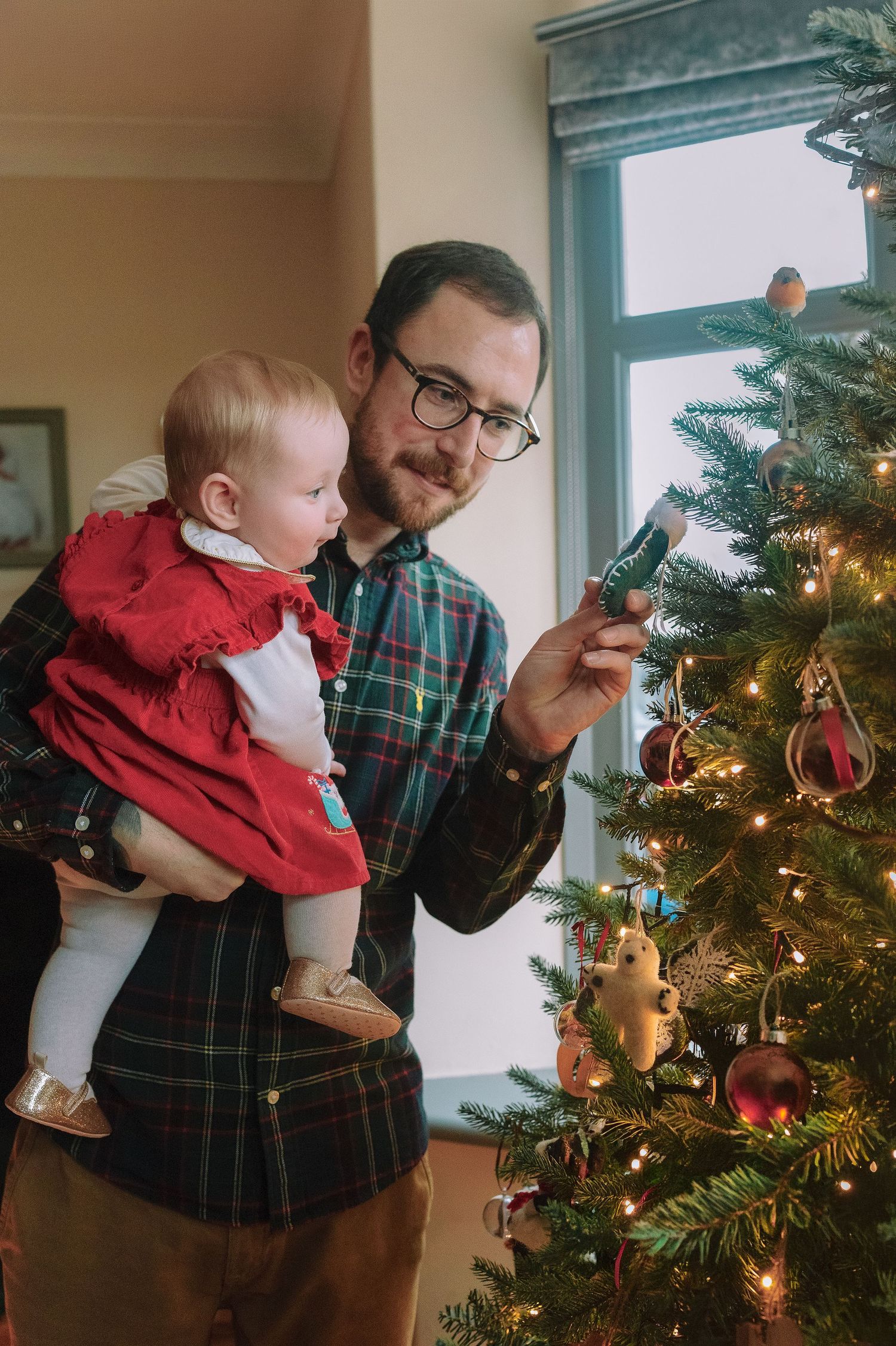 Father holding baby in red dress while placing ornament on decorated Christmas tree indoors.