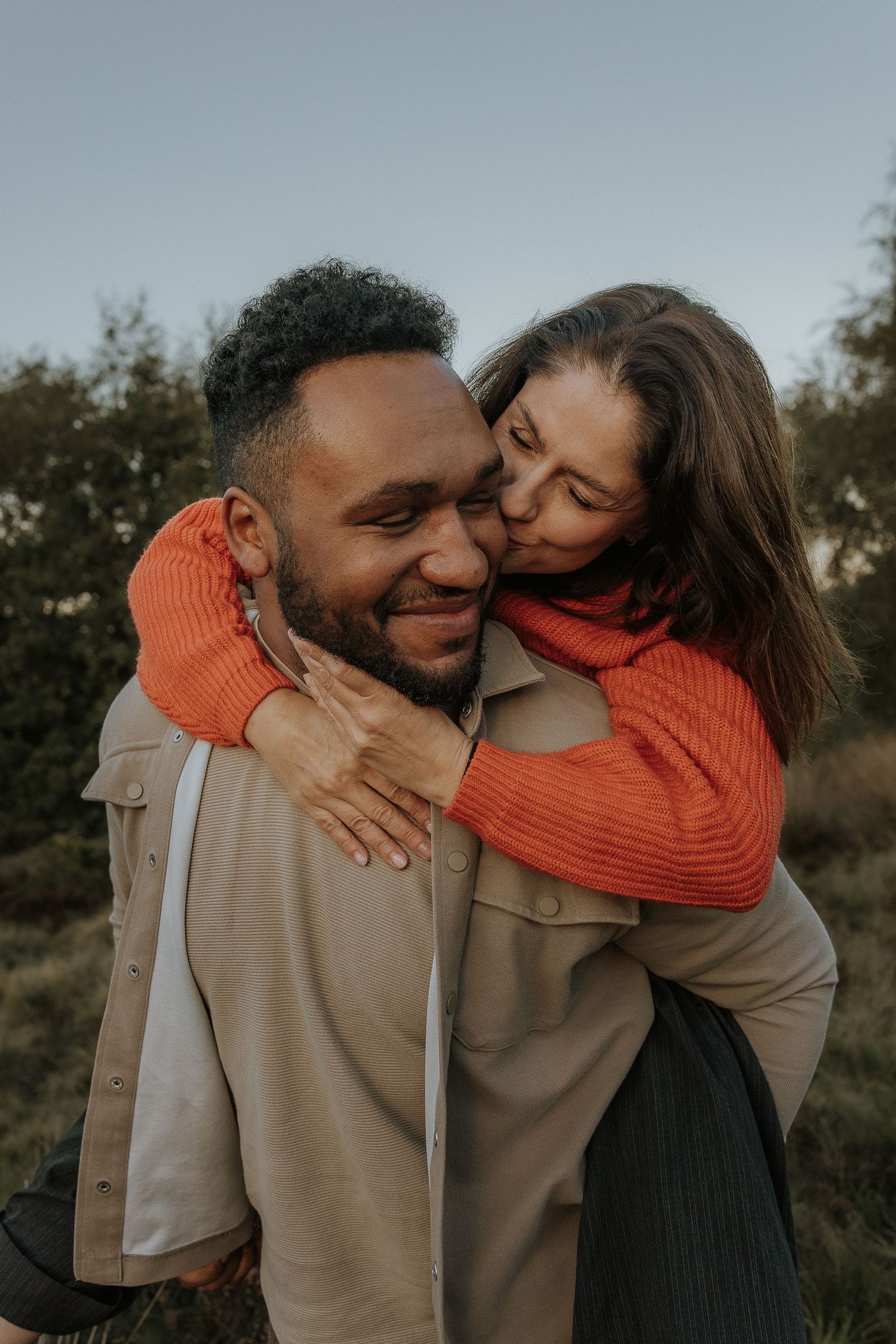 A couple shares a tender moment outdoors during a sunset photoshoot wearing orange and beige clothing.