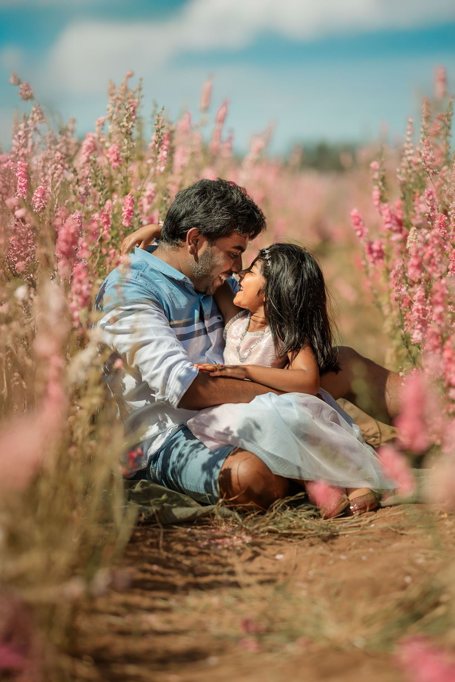 A man and young girl share a tender moment sitting in a pink wildflower field on a sunny day.