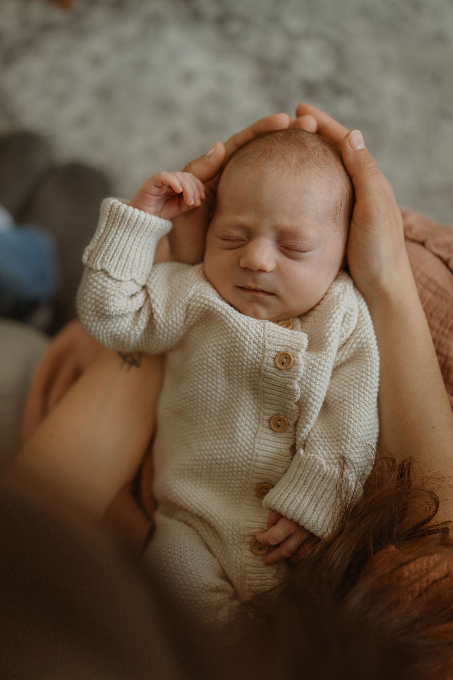 A sleeping newborn baby wearing a white knit cardigan lies peacefully on a brown blanket.