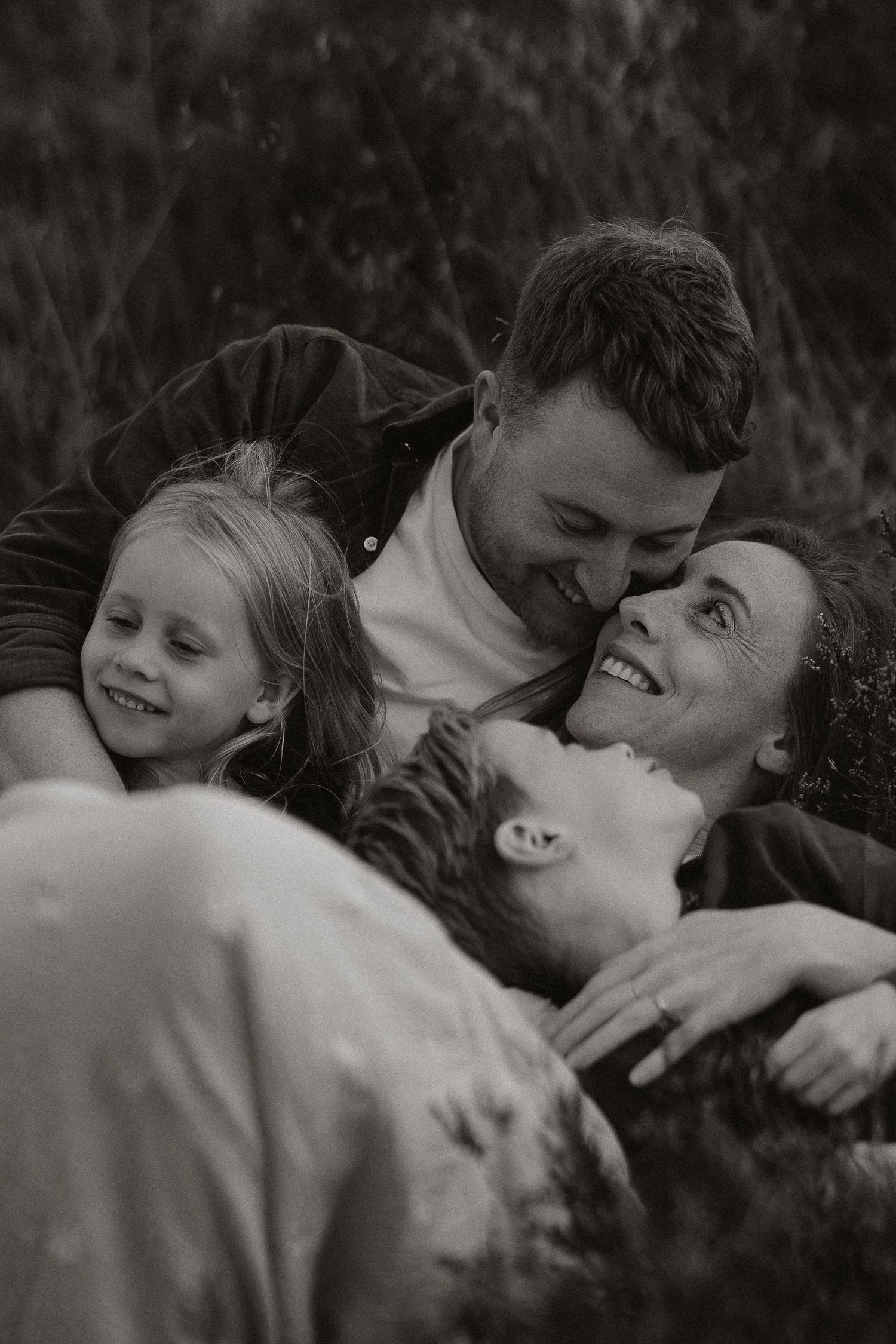A joyful black and white portrait of a family lying in the grass together, sharing intimate laughter and embraces.