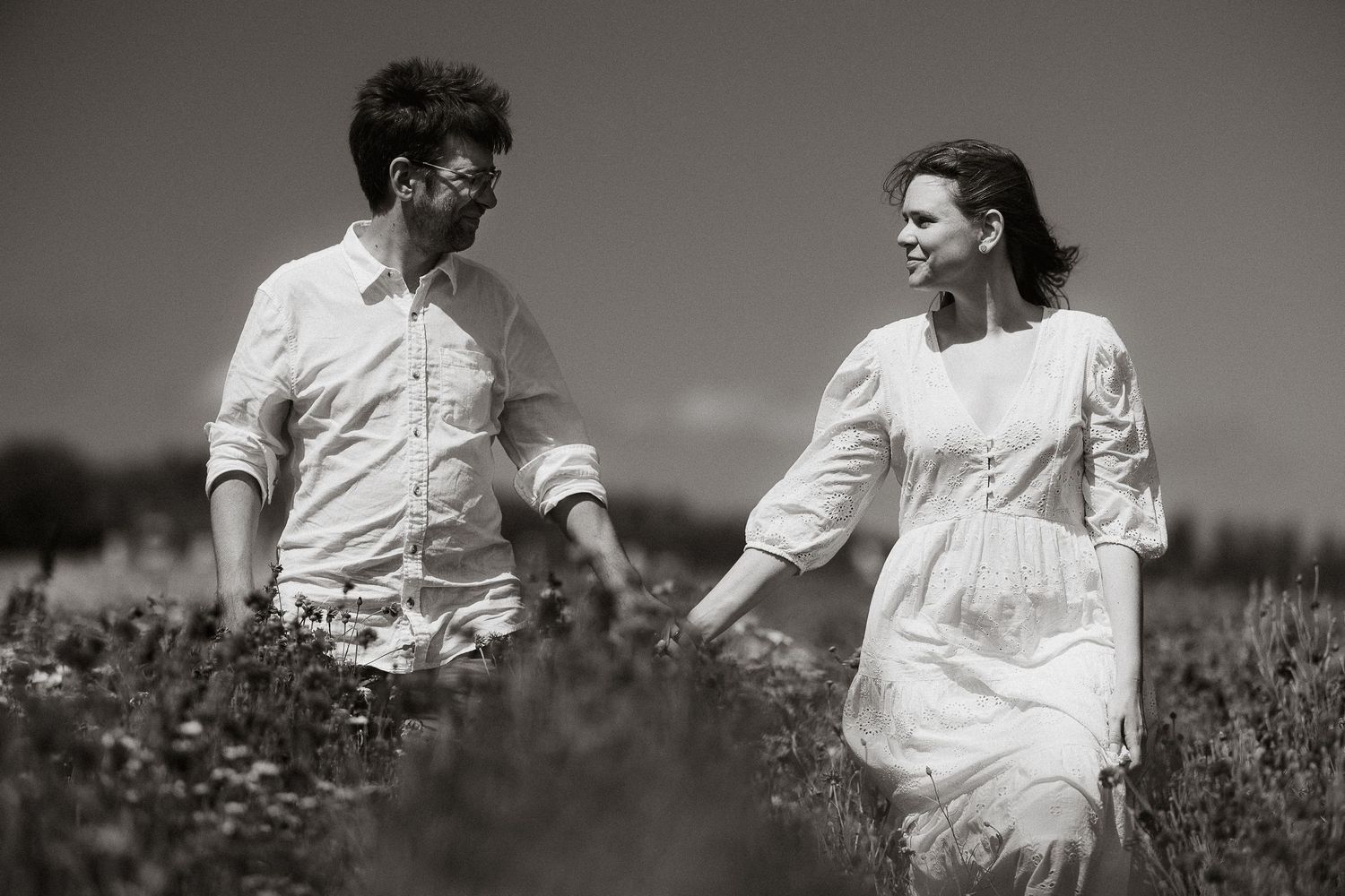 A couple walks hand in hand through a sunny wildflower field, smiling at each other in a romantic black and white photo.