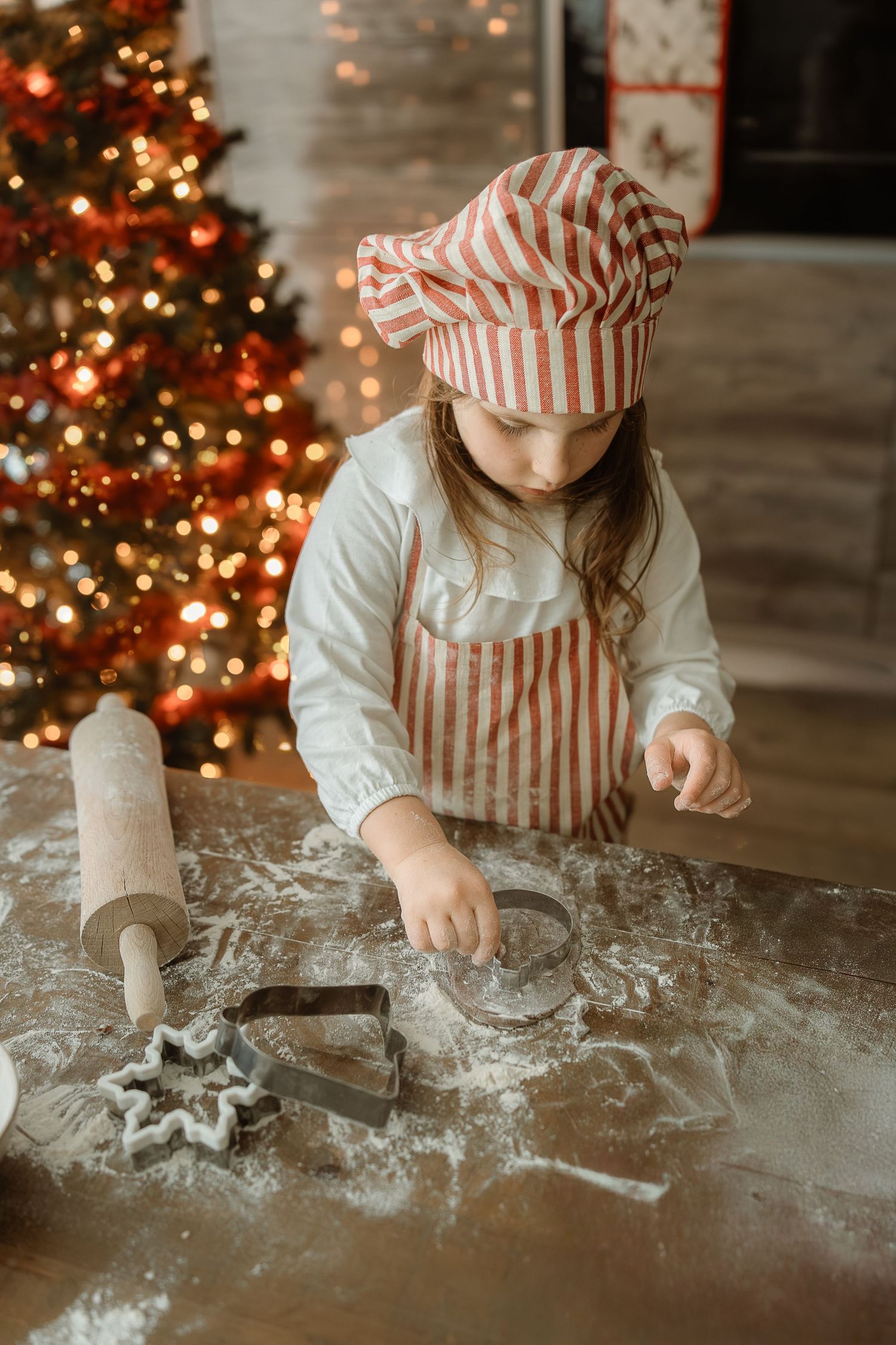 Young girl in striped chef hat and apron cutting Christmas cookies with cookie cutters near decorated tree.