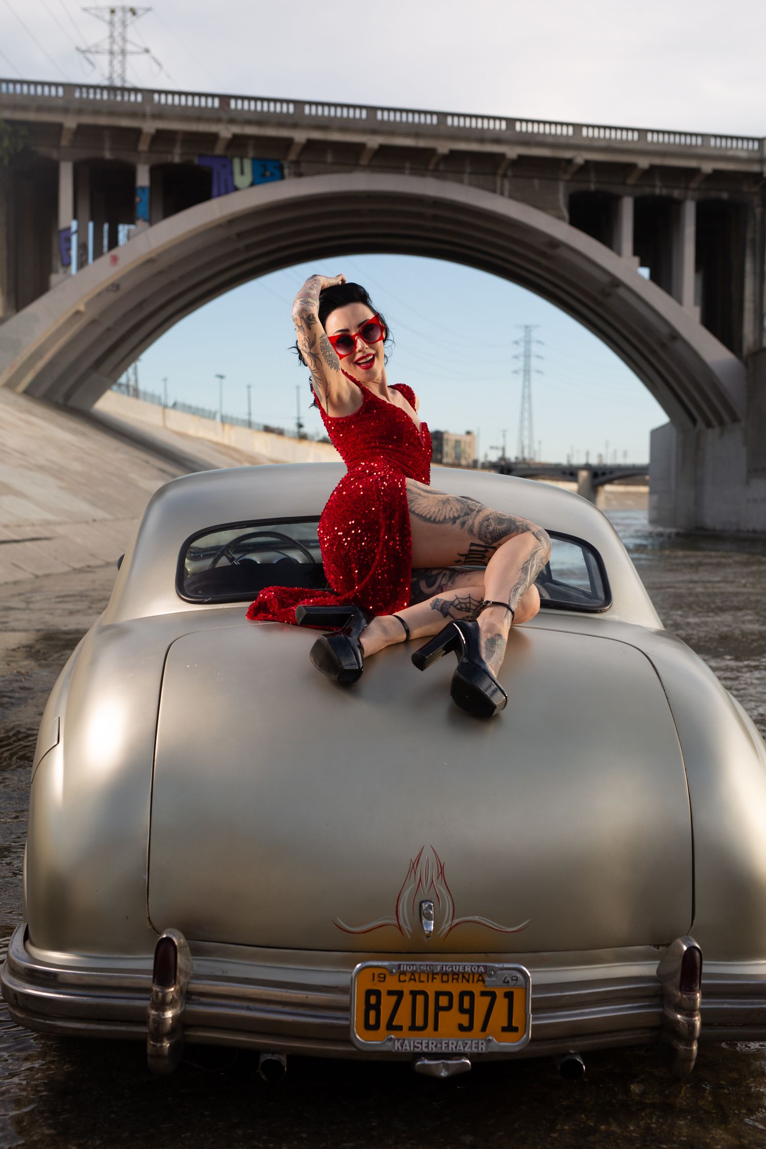 A vintage silver car parked under a concrete bridge arch with someone in a sparkly red dress posing on the trunk.