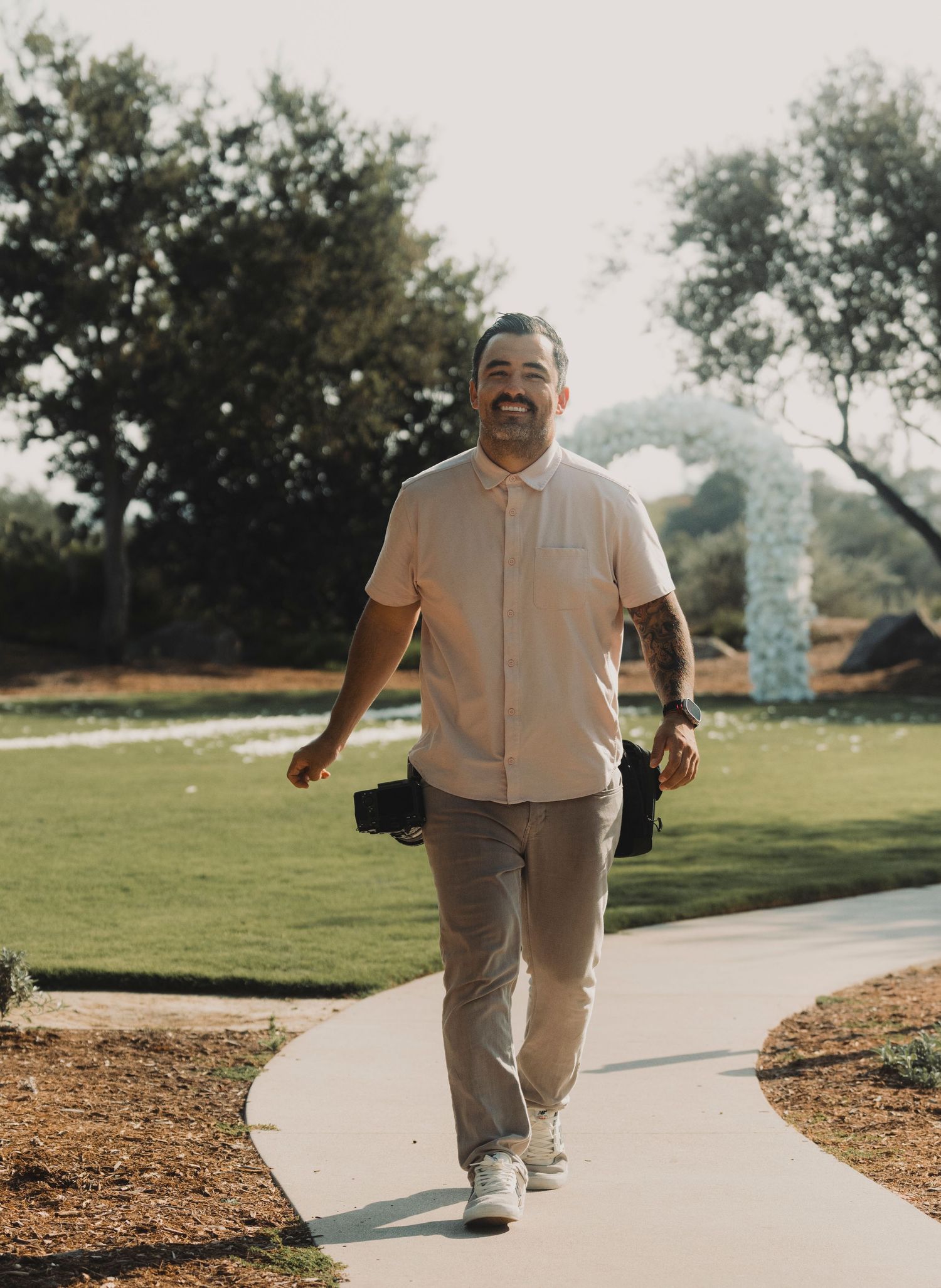 Laughing tattooed photographer in pink shirt carrying cameras while walking along sunny outdoor venue path.