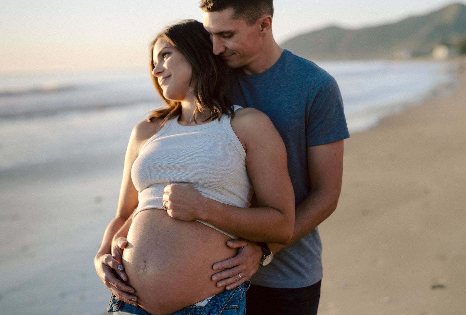 Man embracing pregnant partner from behind on sandy beach at dusk.