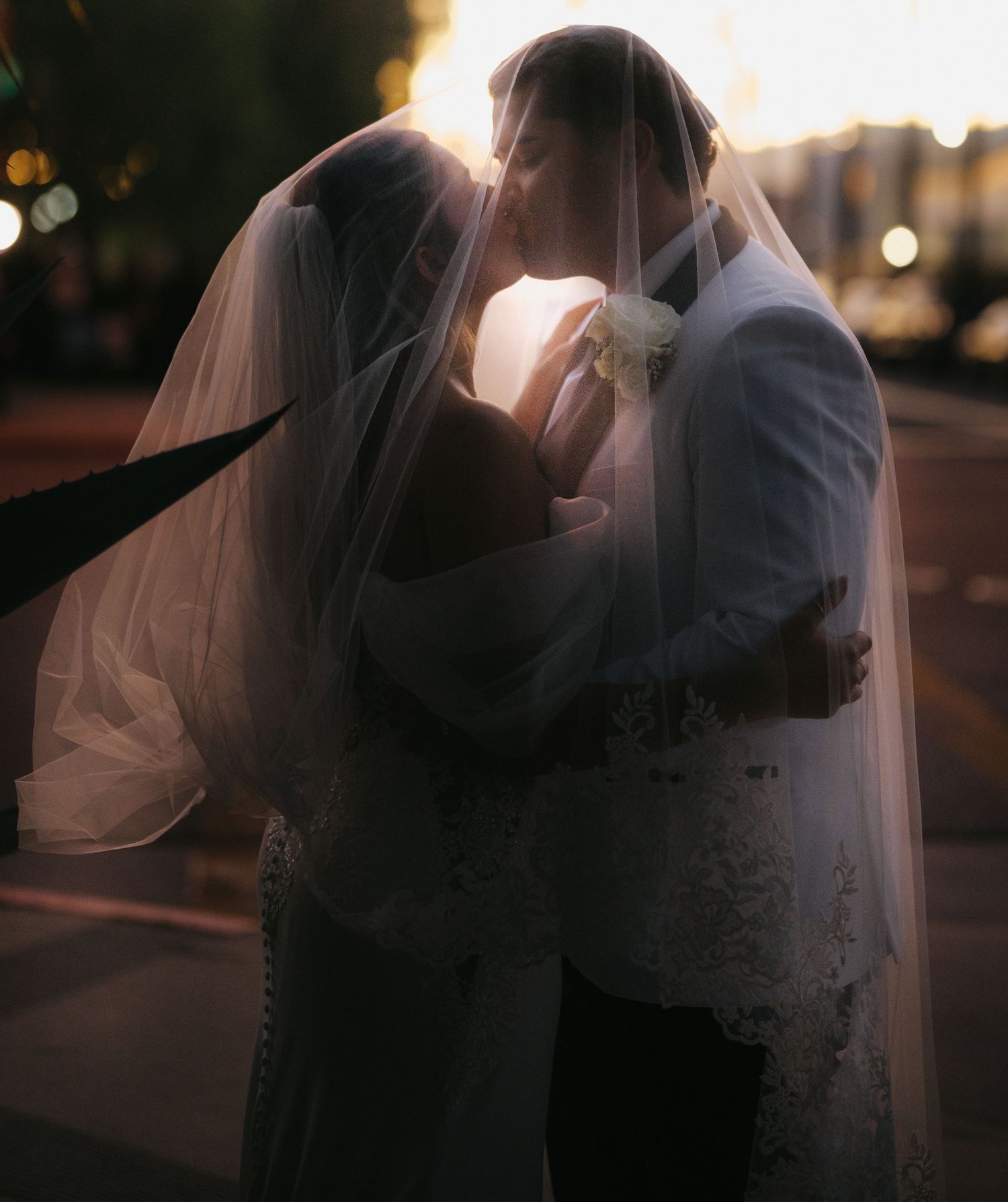 Bride and groom share a romantic kiss under a flowing veil at golden hour sunset.