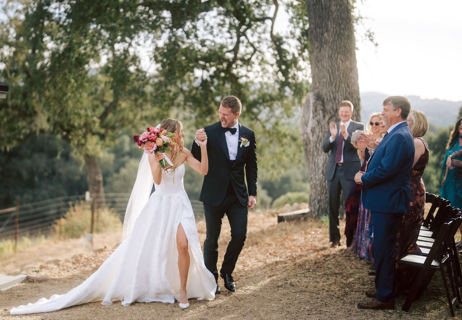 Joyful bride and groom walk back down aisle after outdoor wedding ceremony under oak tree.