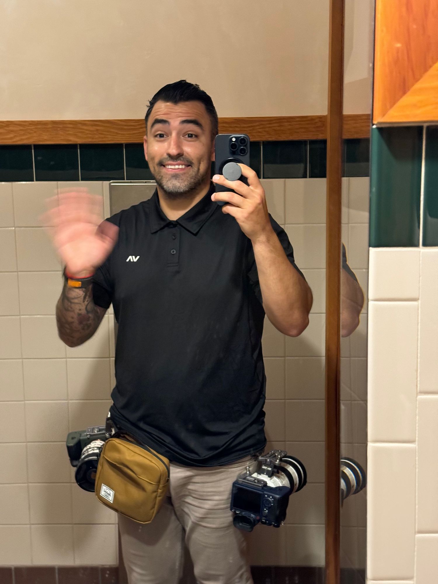 Man taking mirror selfie in bathroom holding cameras with tattoos visible on arm.