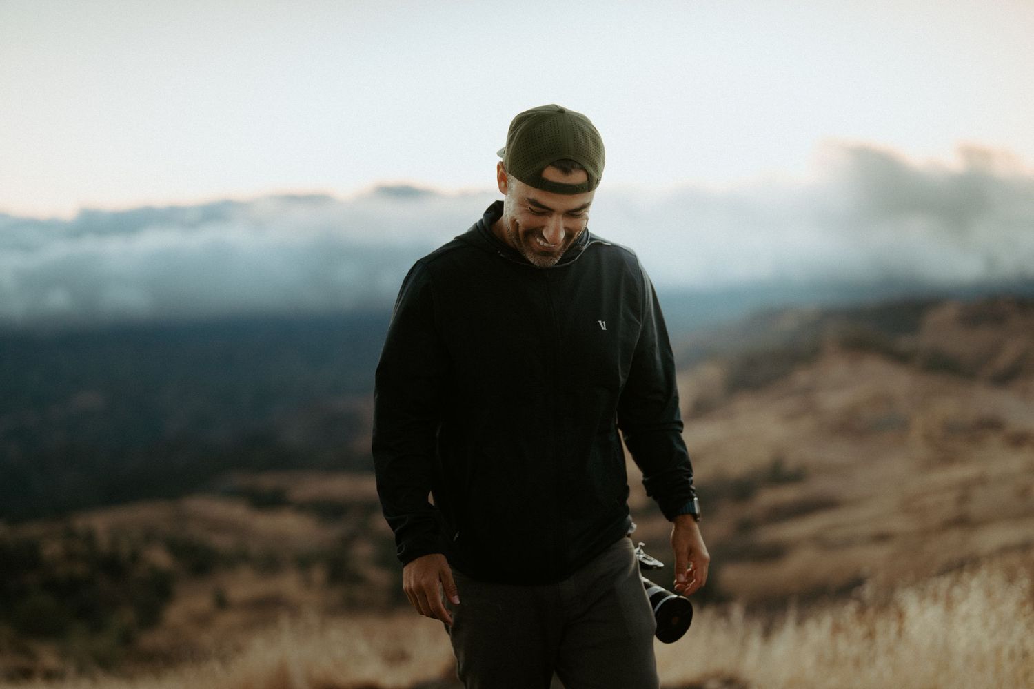 Photographer in black hoodie holding camera on misty mountain hillside at dusk.