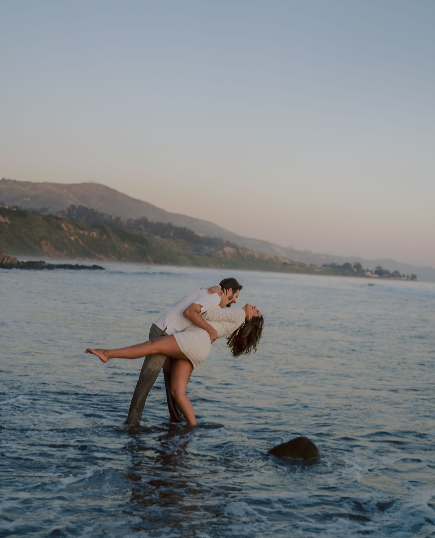 Man lifting woman in joyful embrace while standing in ocean waves at sunset beach.
