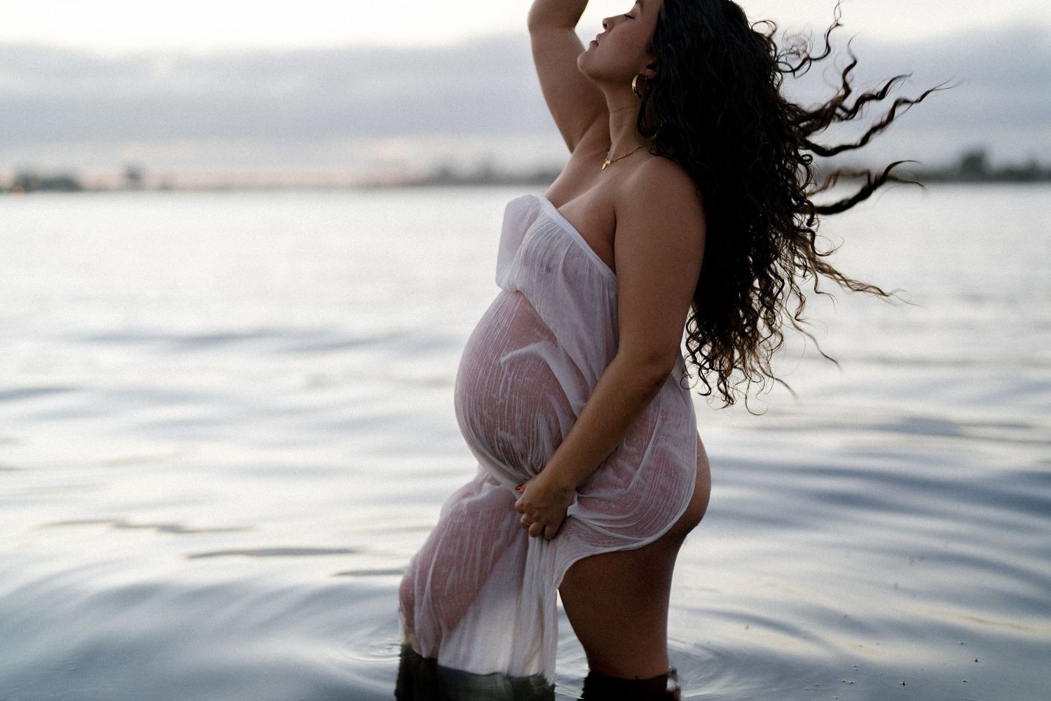 Pregnant woman in sheer white wrap standing in water at dusk with wind-blown curly hair.