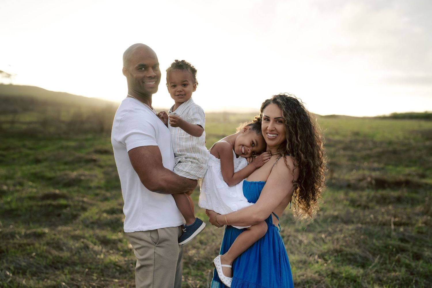 Happy family of four with two babies posing in sunlit green field at sunset.