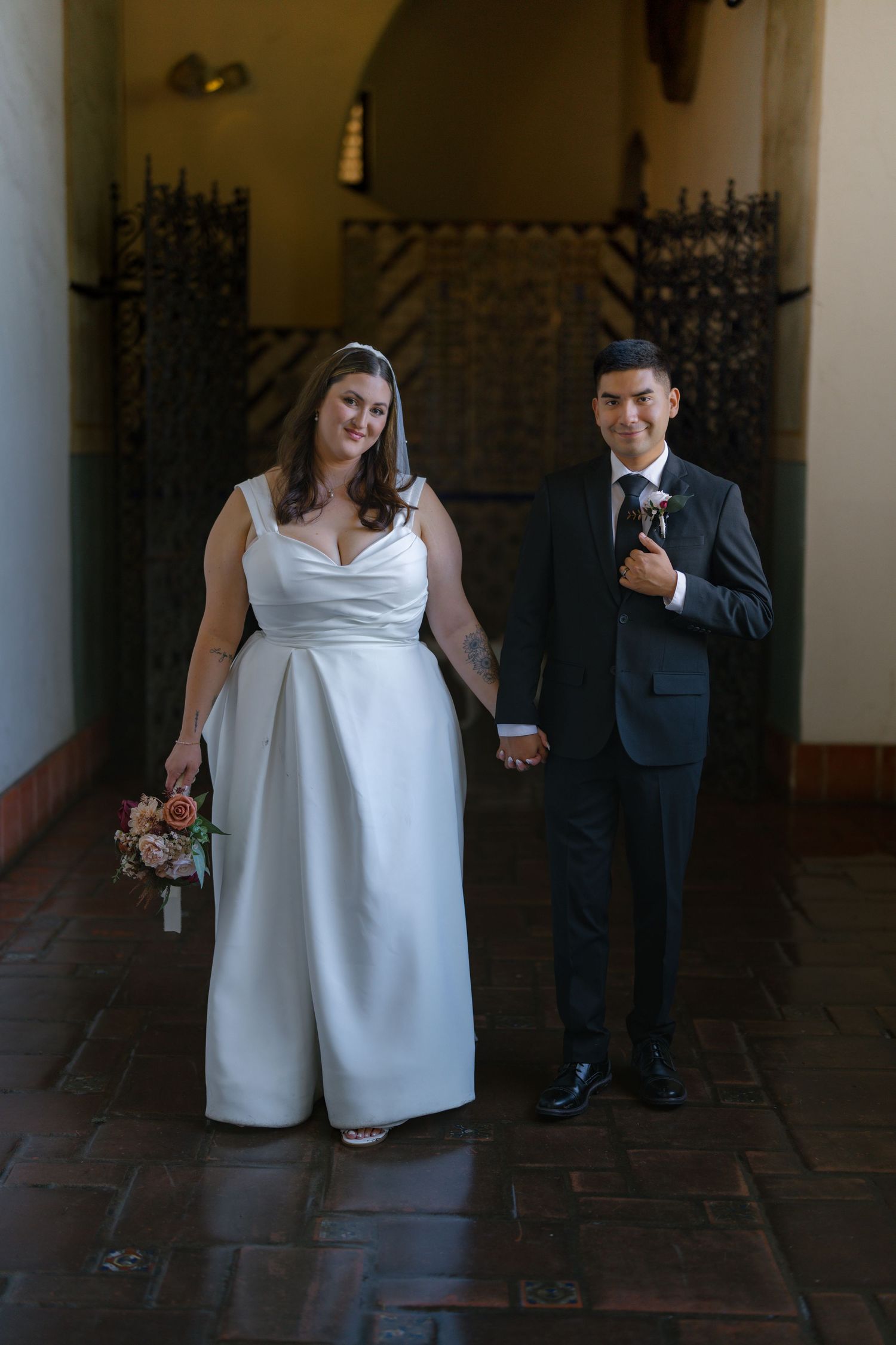 Smiling bride in sleek white gown holds bouquet while groom in dark suit holds her hand indoors.