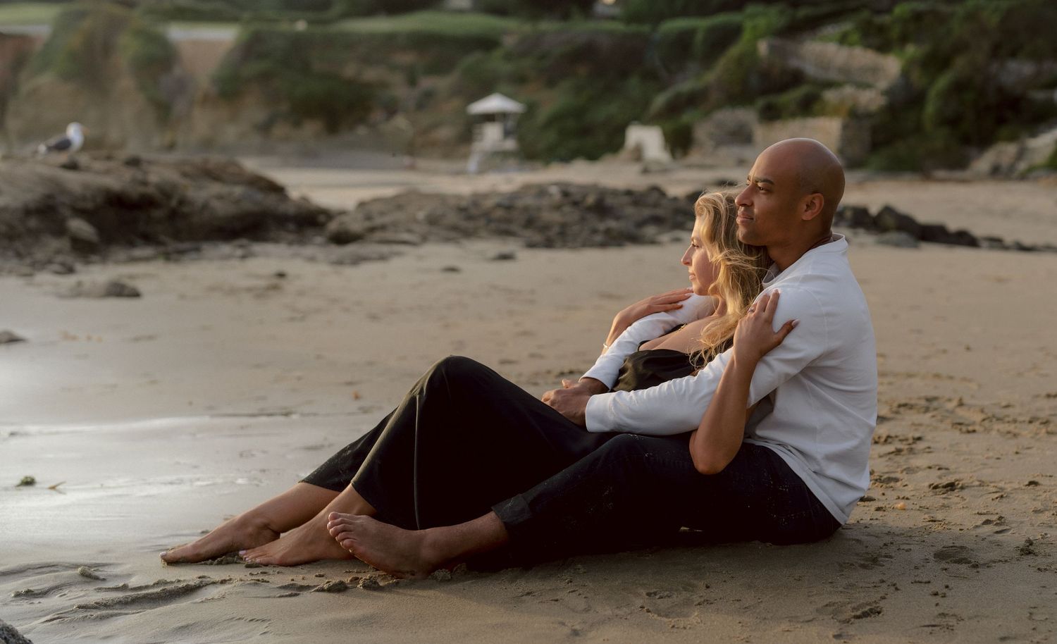 Couple sitting together on a sandy beach at dusk, sharing a tender romantic moment by the ocean.