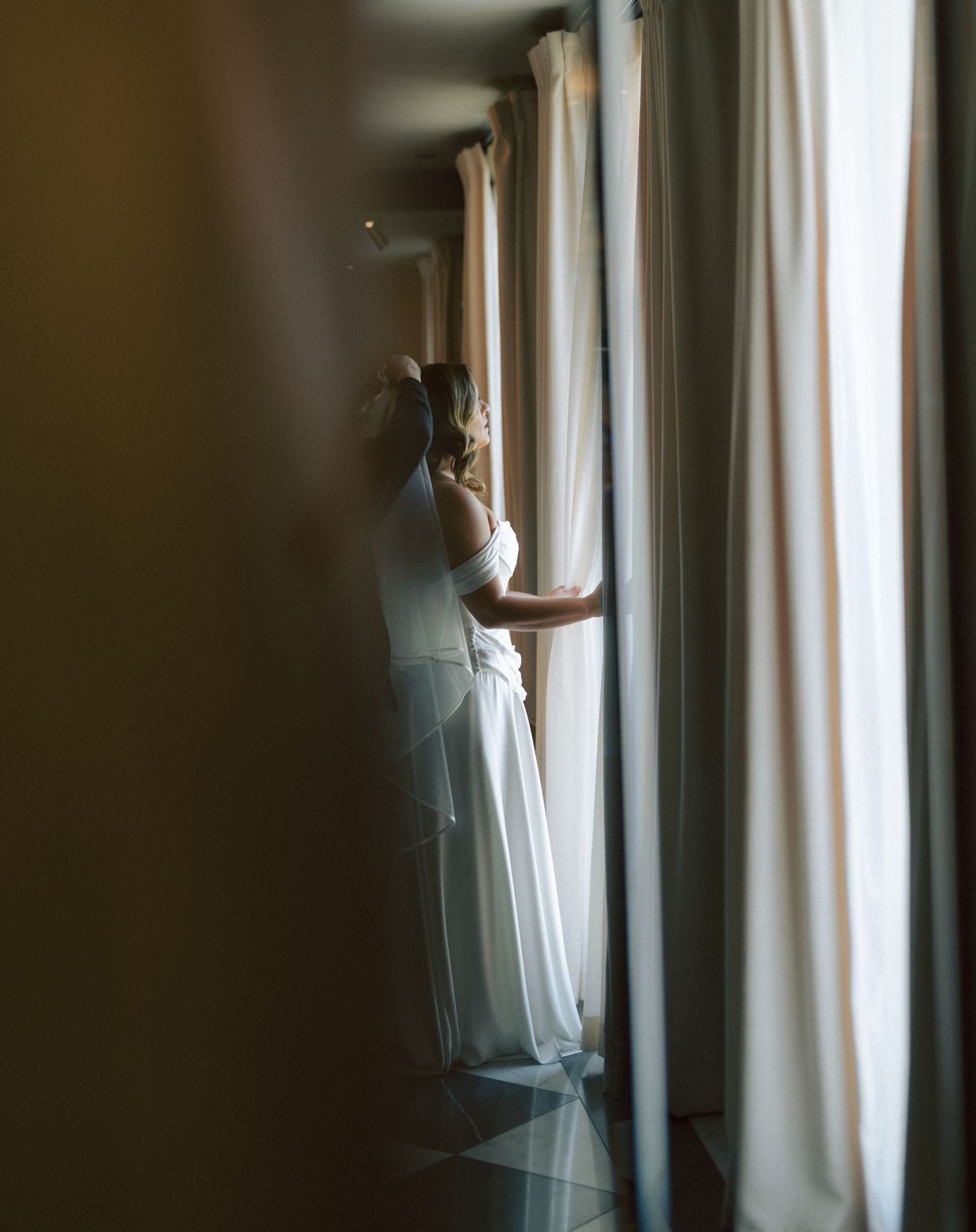 Bride in white gown gazing through curtained window in elegant room.