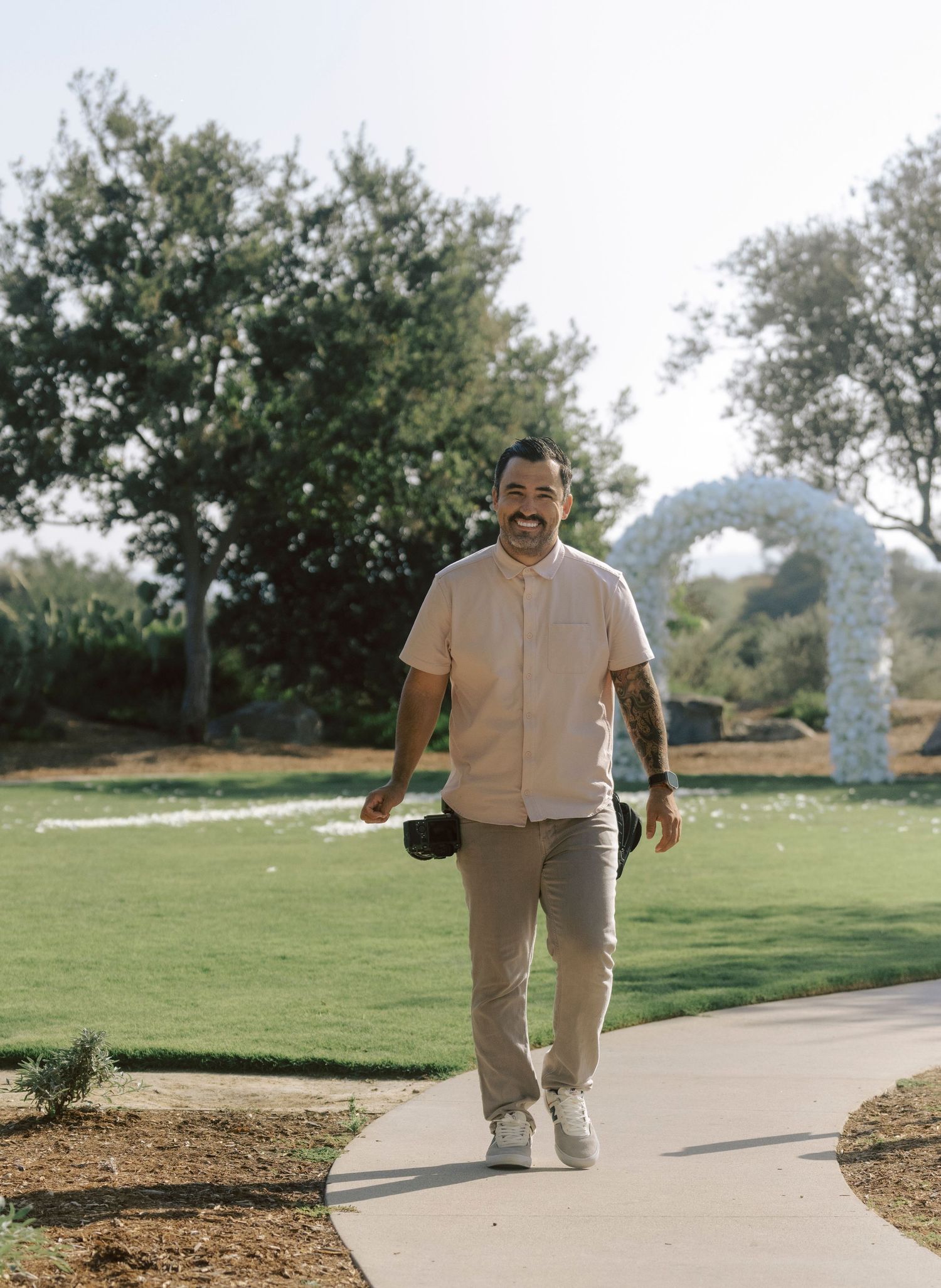 Smiling tattooed photographer in pink shirt walking along garden path carrying camera at outdoor venue.