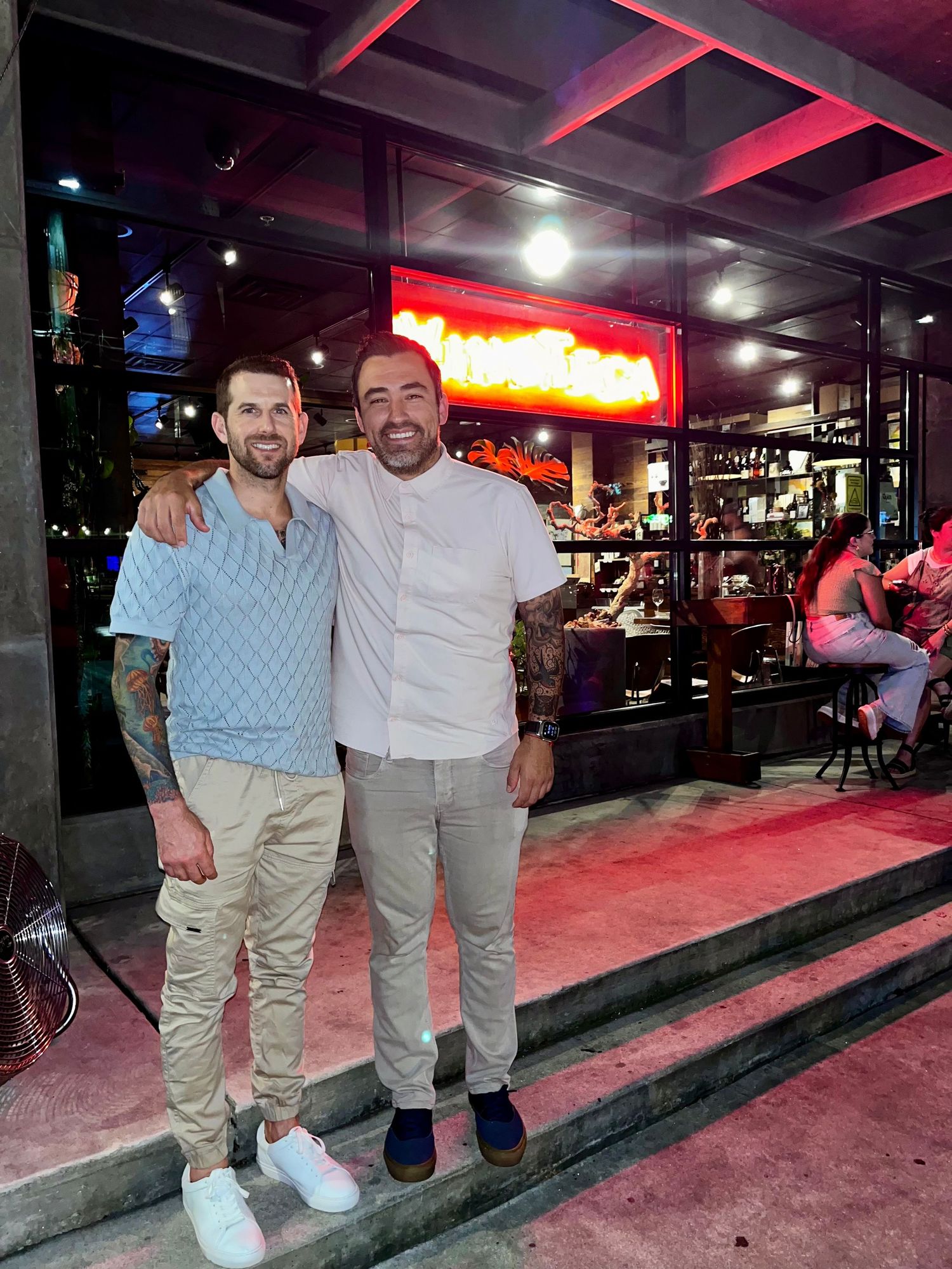 Two men smiling and posing together outside a restaurant with neon signage at night.