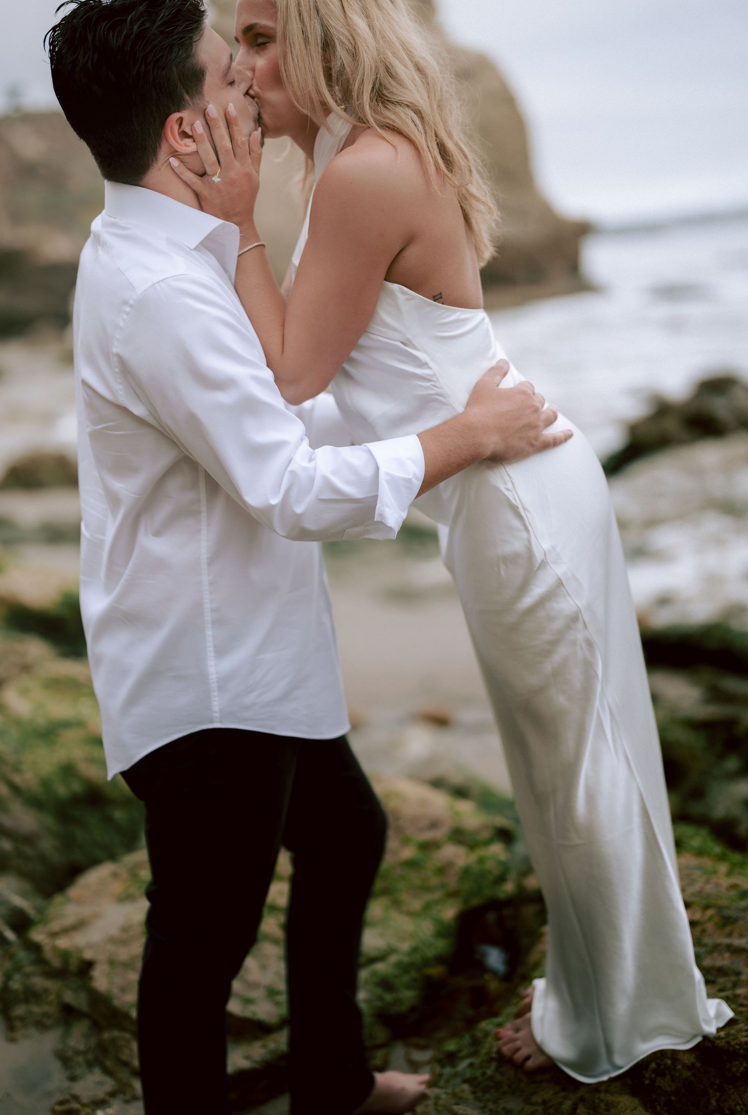 Couple sharing a passionate kiss on rocky beach, woman in white dress embraced by man in white shirt.