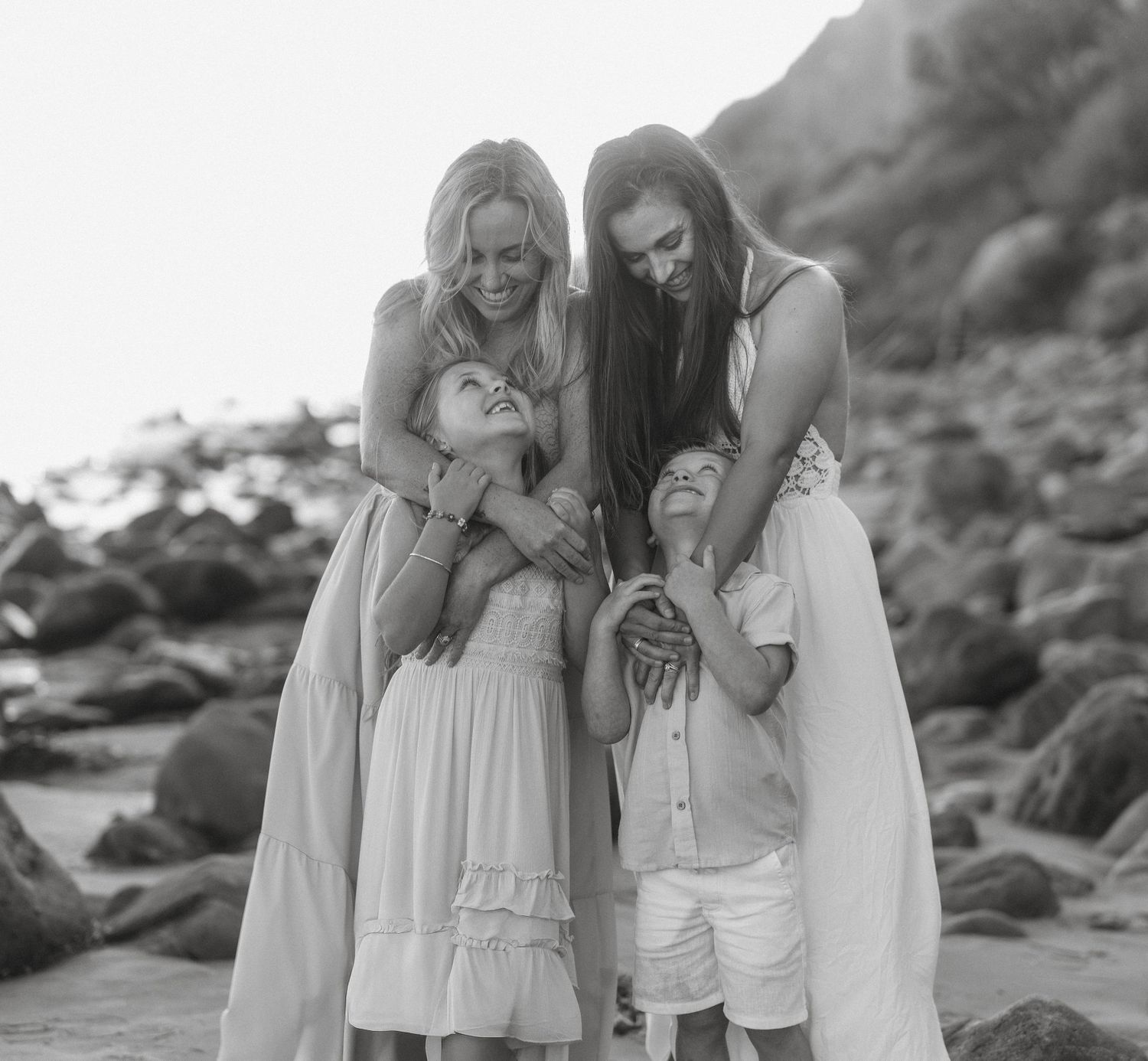 Black and white photo of two mothers lovingly embracing their children on rocky beach.