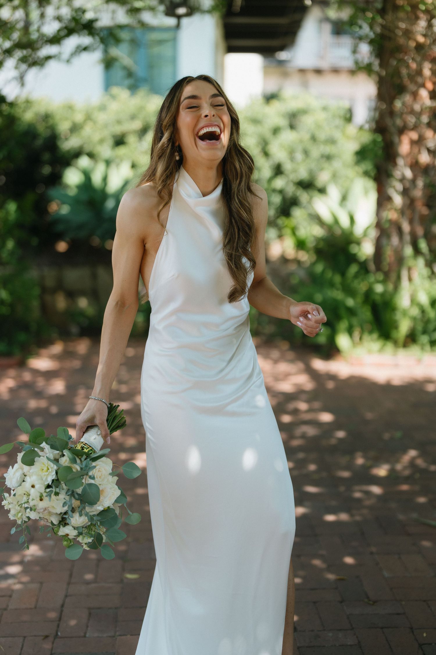 Joyful bride in white halter gown laughing while holding white floral bouquet on brick path.