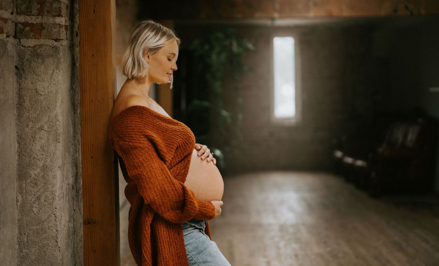 Pregnant blonde woman in orange knit sweater leaning against wall, cradling baby bump in rustic indoor space.