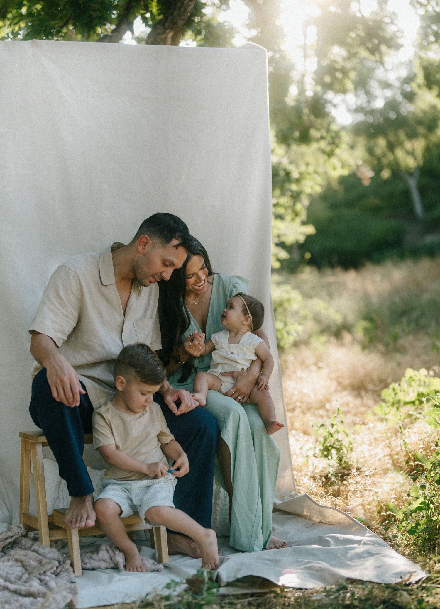 Family of four sharing a tender moment seated outdoors against a neutral linen backdrop.