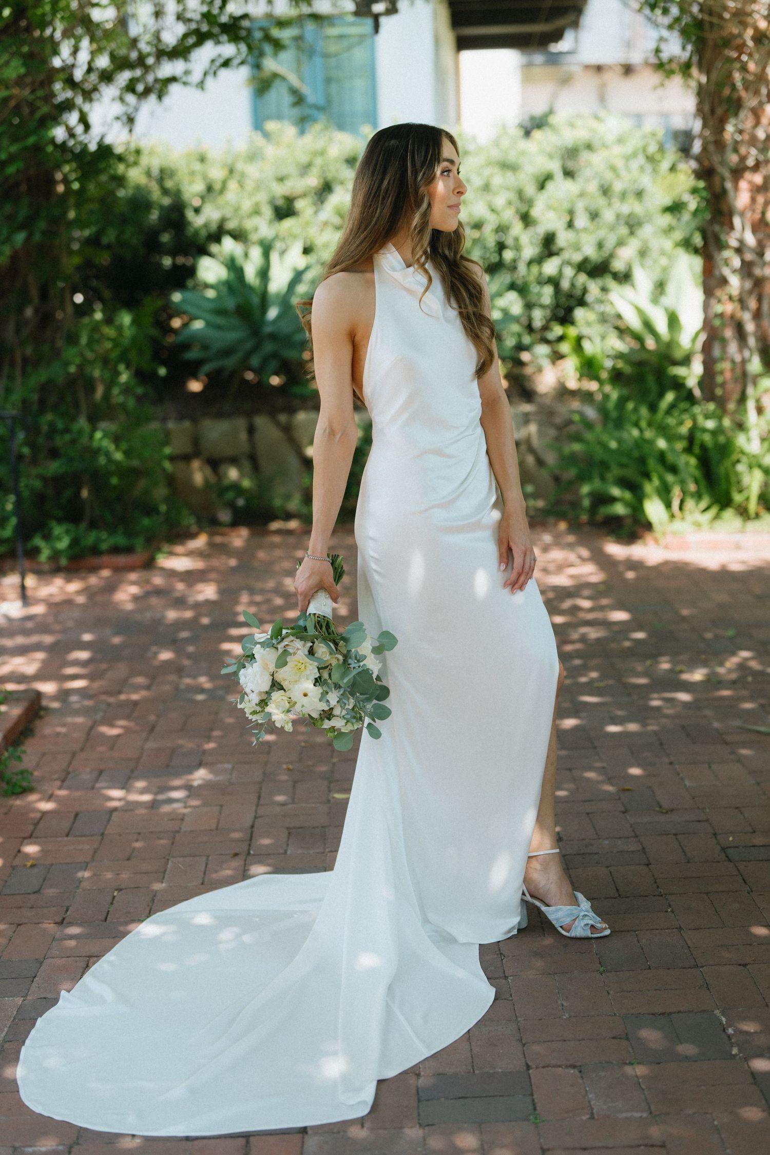Elegant bride in white halter wedding gown with train holds bouquet on sunlit brick path.
