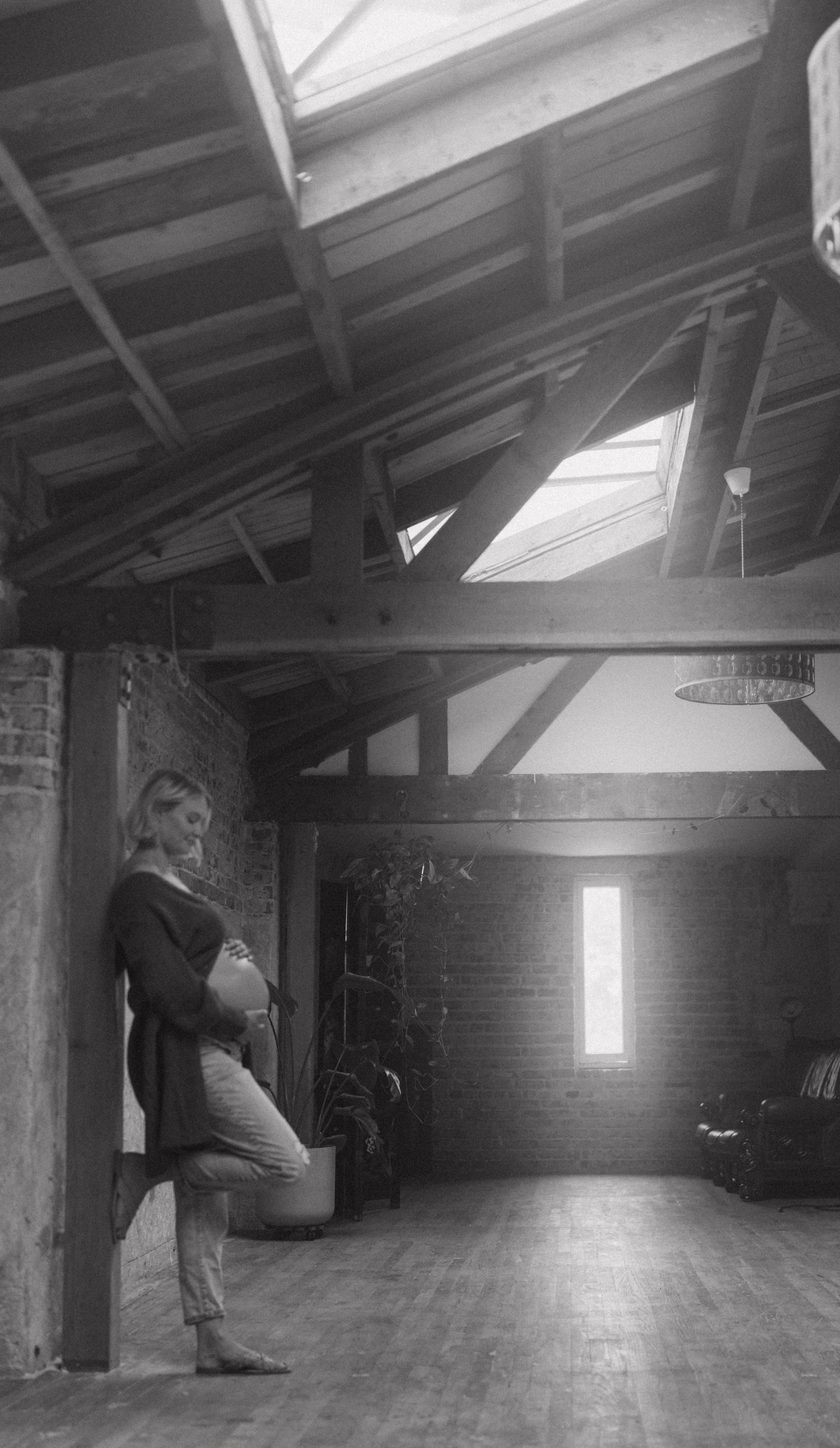 Black and white photo of pregnant woman leaning against brick wall in rustic loft space with skylights.