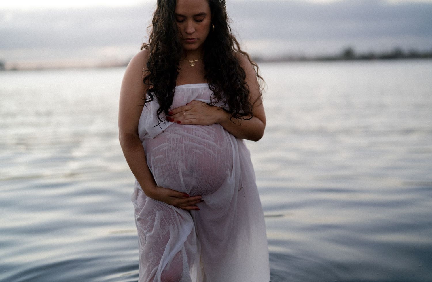 Pregnant woman in flowing white dress cradling baby bump while standing in calm water at sunset.