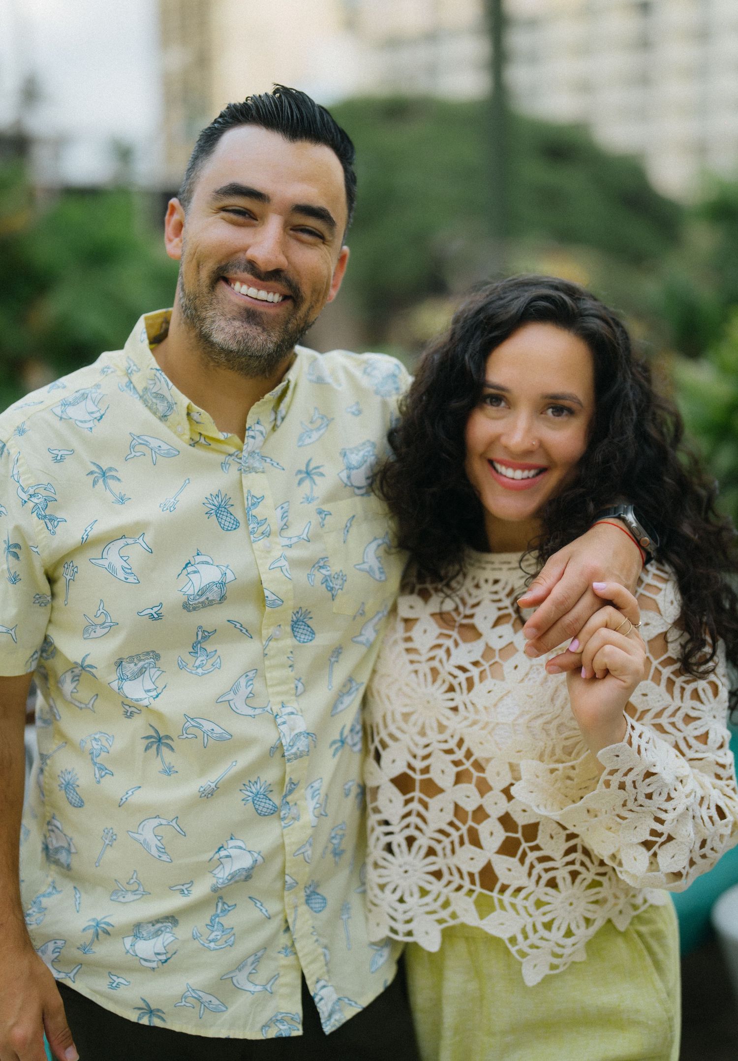 Smiling couple posing together outdoors, man in tropical shirt, woman in lace top.