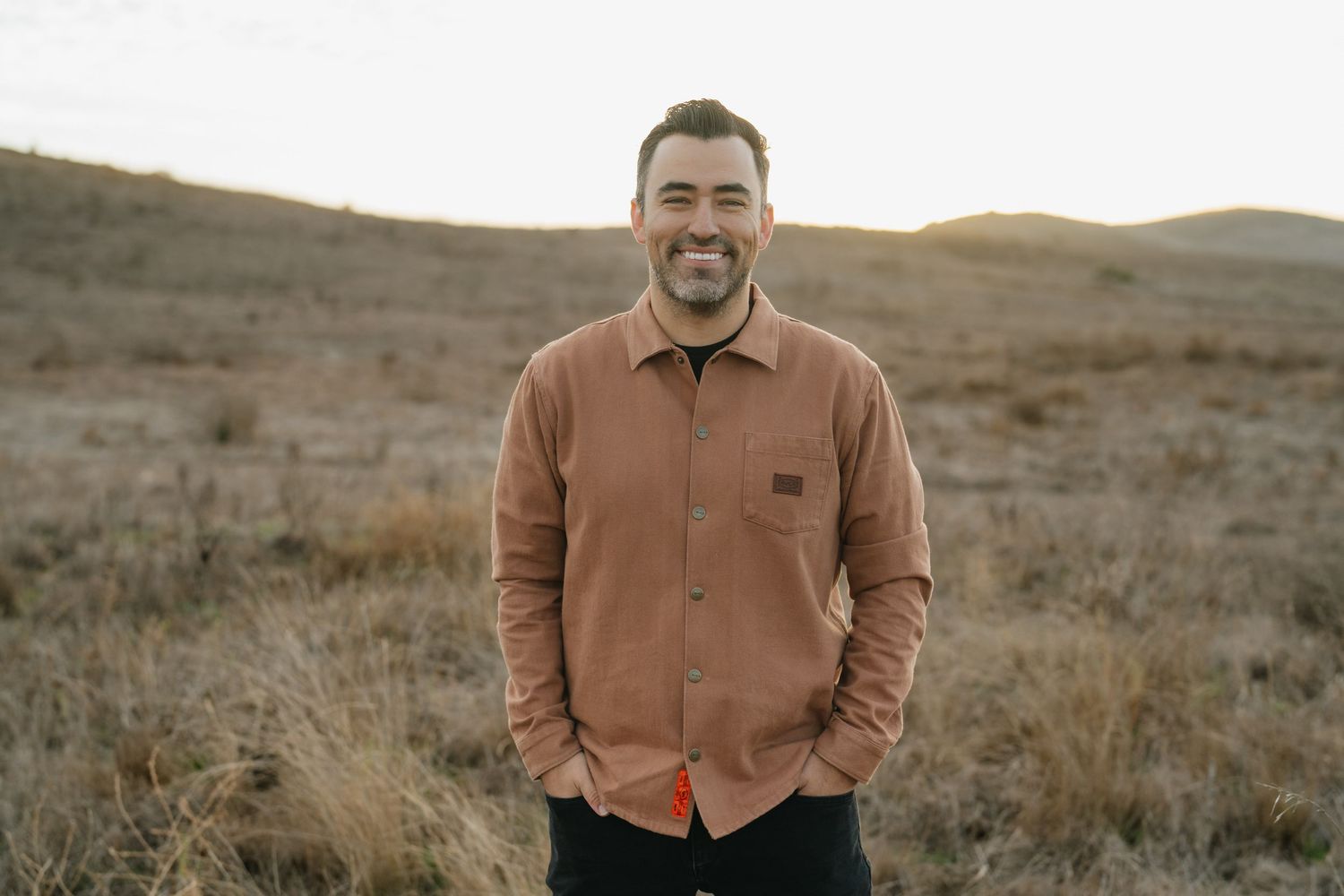 Smiling man in rust shirt standing in golden field at sunset.