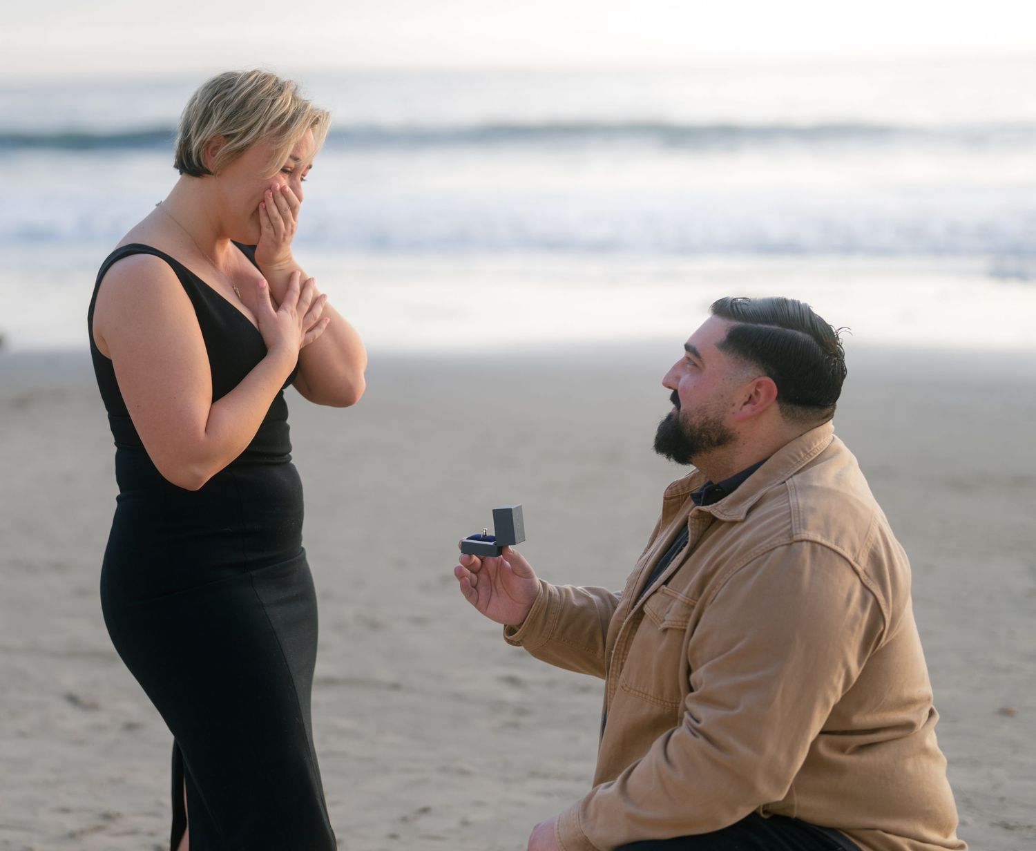 Man proposing on beach at sunset, kneeling with ring box while woman covers her mouth in surprise.