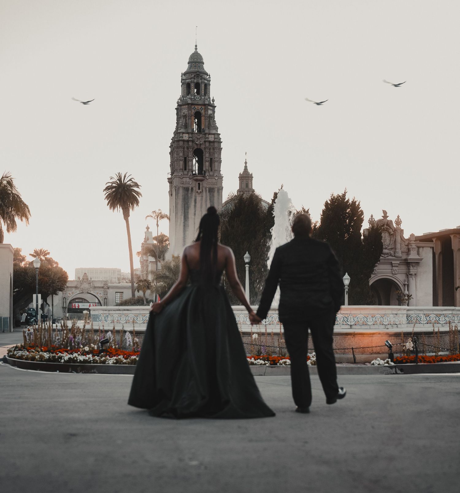 Couple in formal black attire holding hands before a historic tower and garden at dusk.