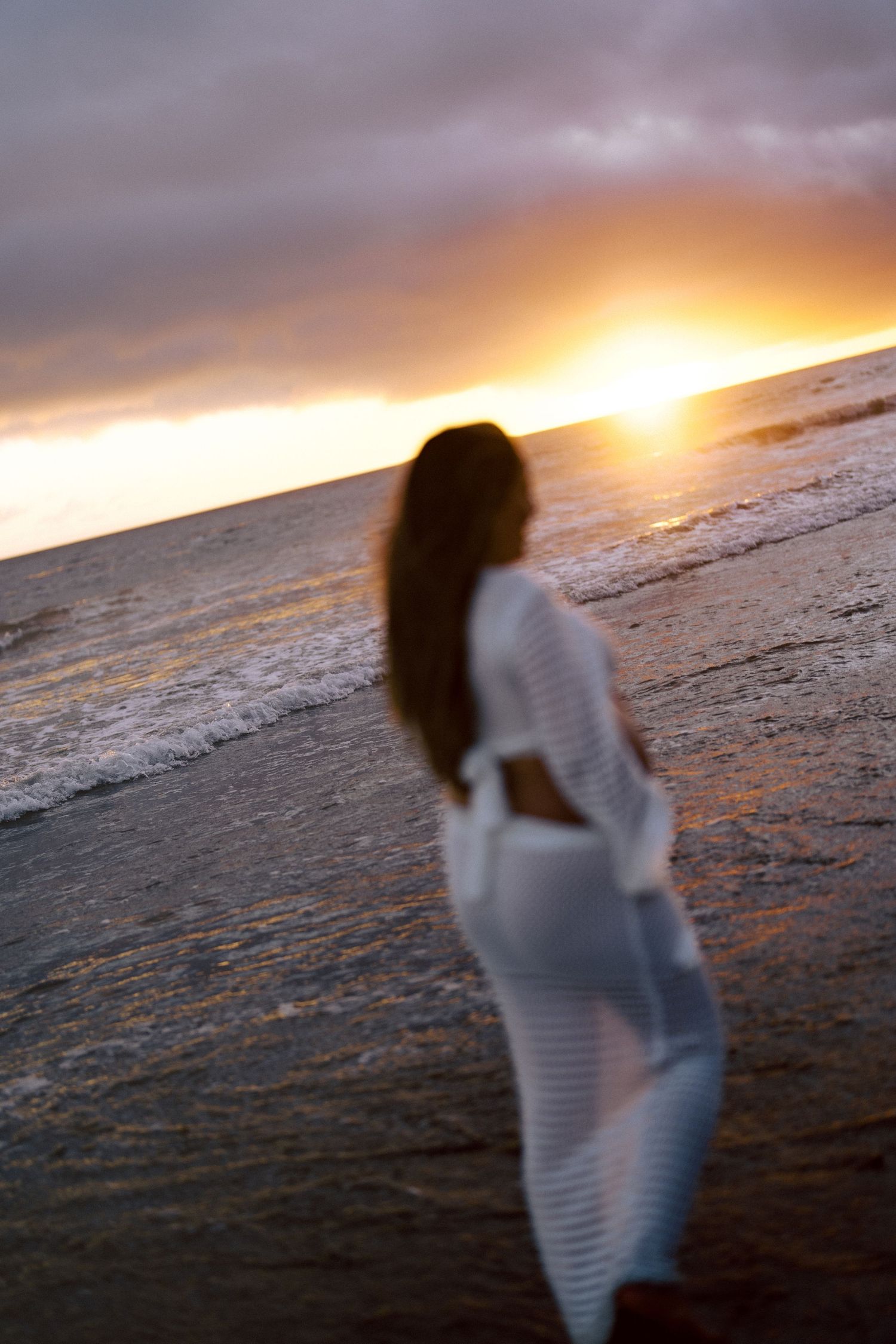 Blurred pregnant woman in white lace dress walking on beach at vivid sunset.