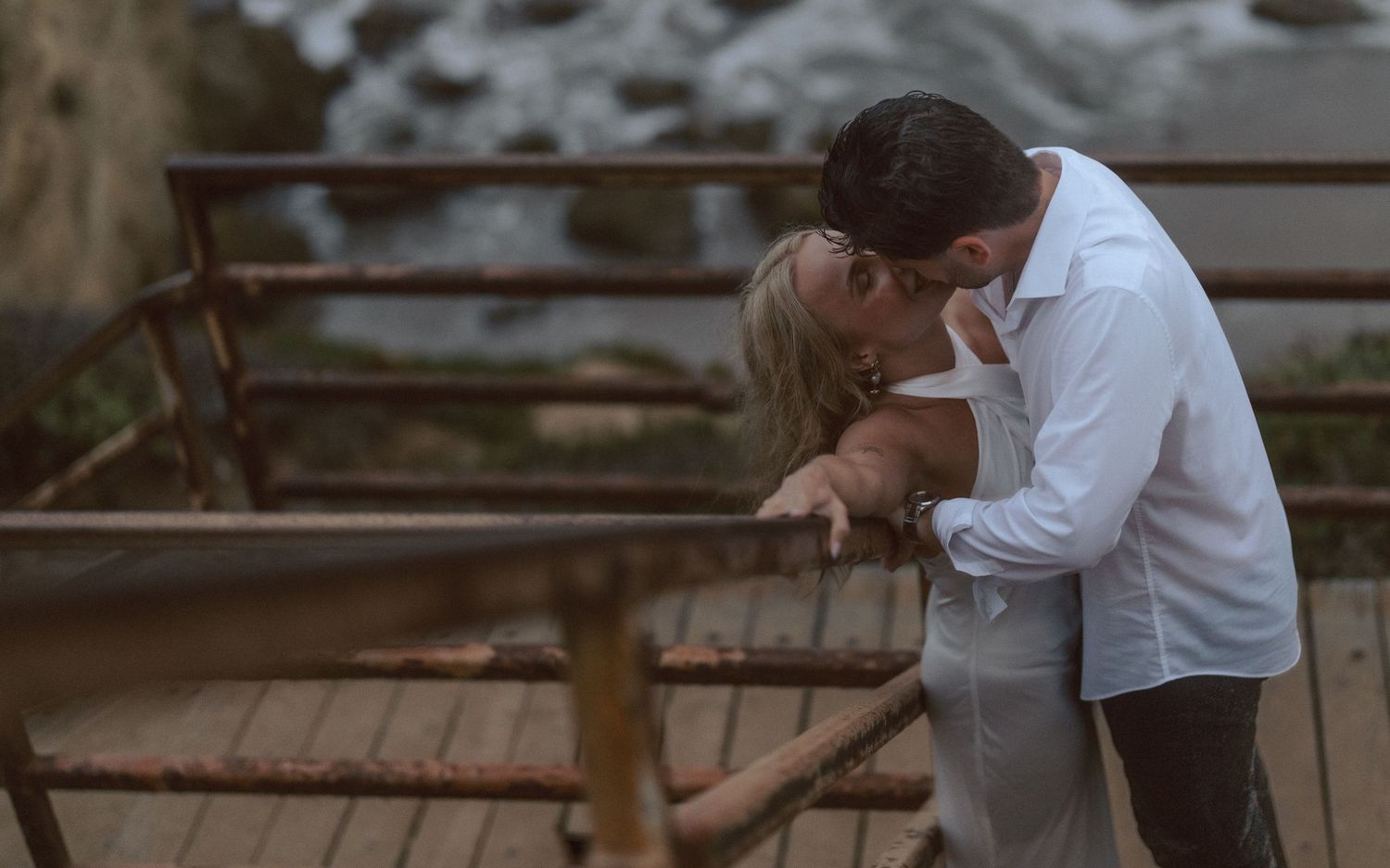 Romantic couple sharing a kiss on a wooden bridge overlooking water outdoors.
