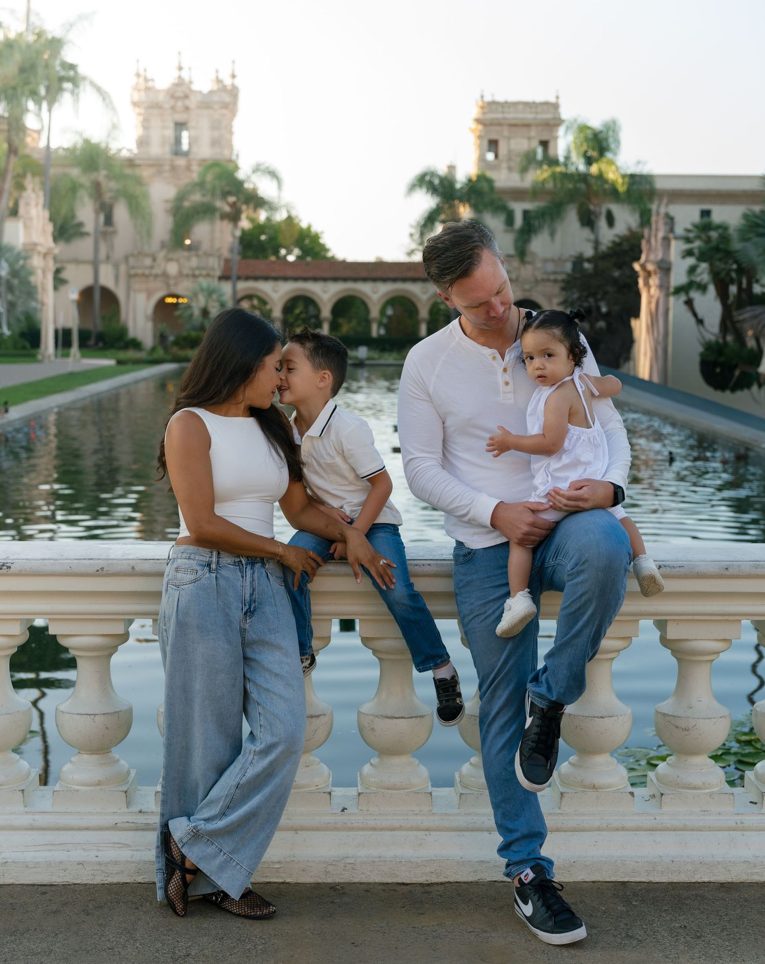 Family of four posing by a fountain at Balboa Park, parents holding young children while smiling warmly.