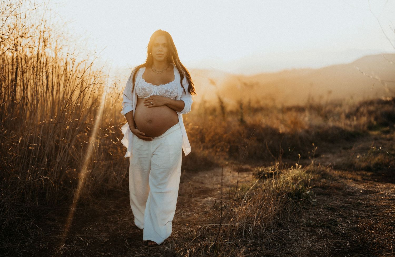 Pregnant woman in flowing white dress walking through a sunlit golden field with mountains in background.