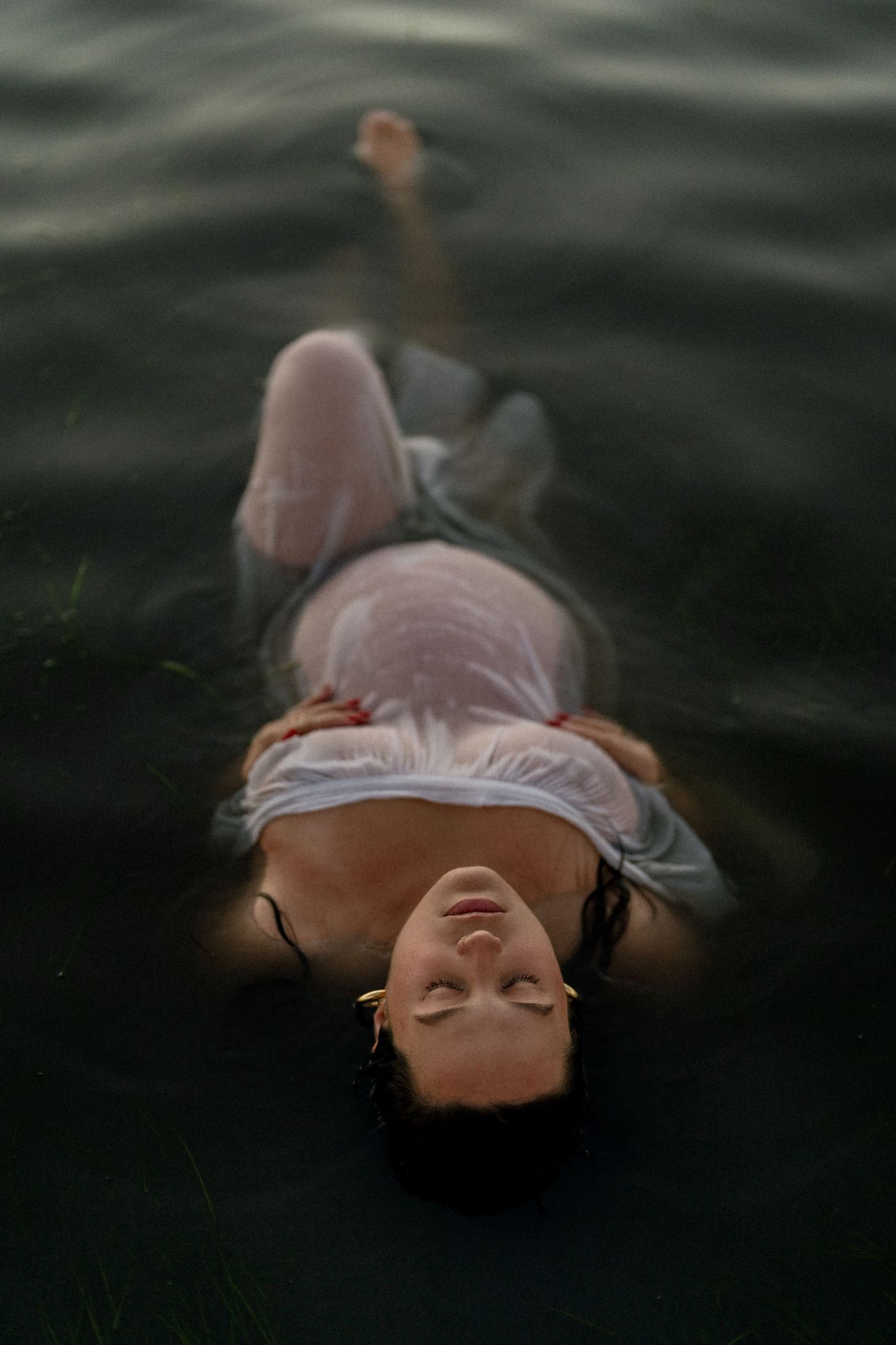 Pregnant woman in wet white dress floats peacefully in dark water for maternity shoot.