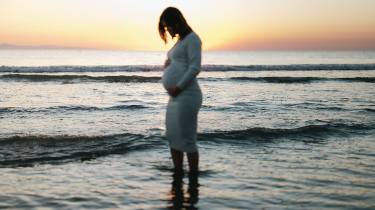 Pregnant woman in white dress standing in ocean waves at colorful sunset.