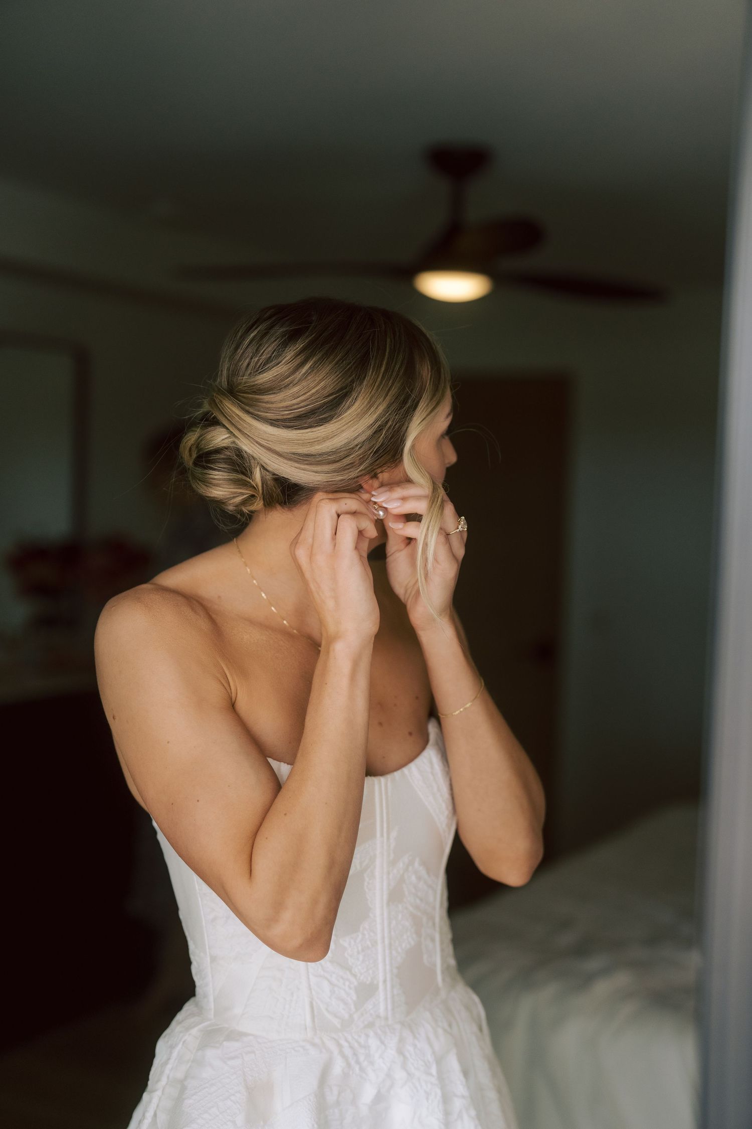 Blonde bride in strapless wedding gown putting on earrings with elegant updo hairstyle in hotel room.