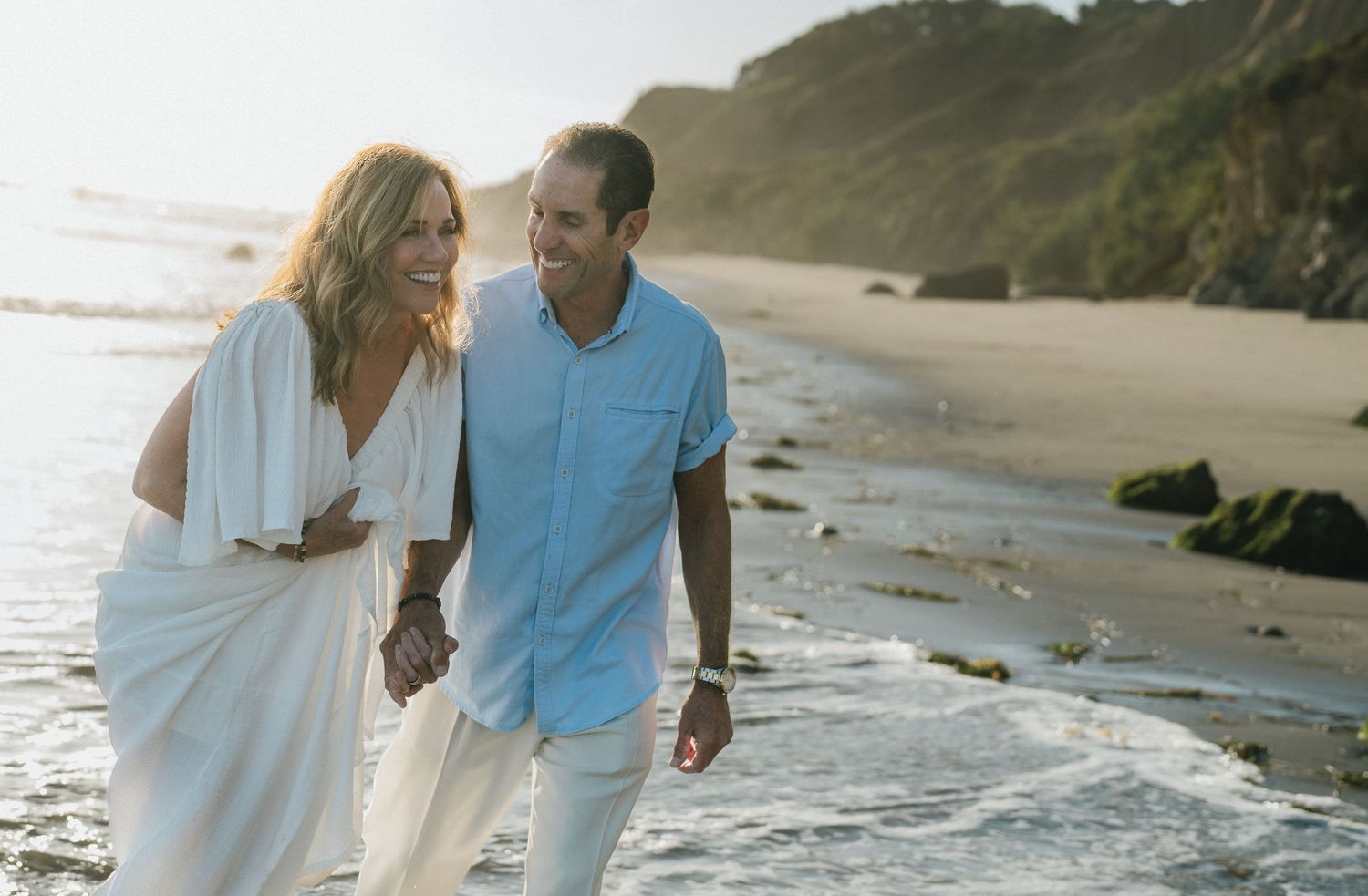Happy couple walking hand-in-hand along a sunny beach with rocky cliffs in the background.