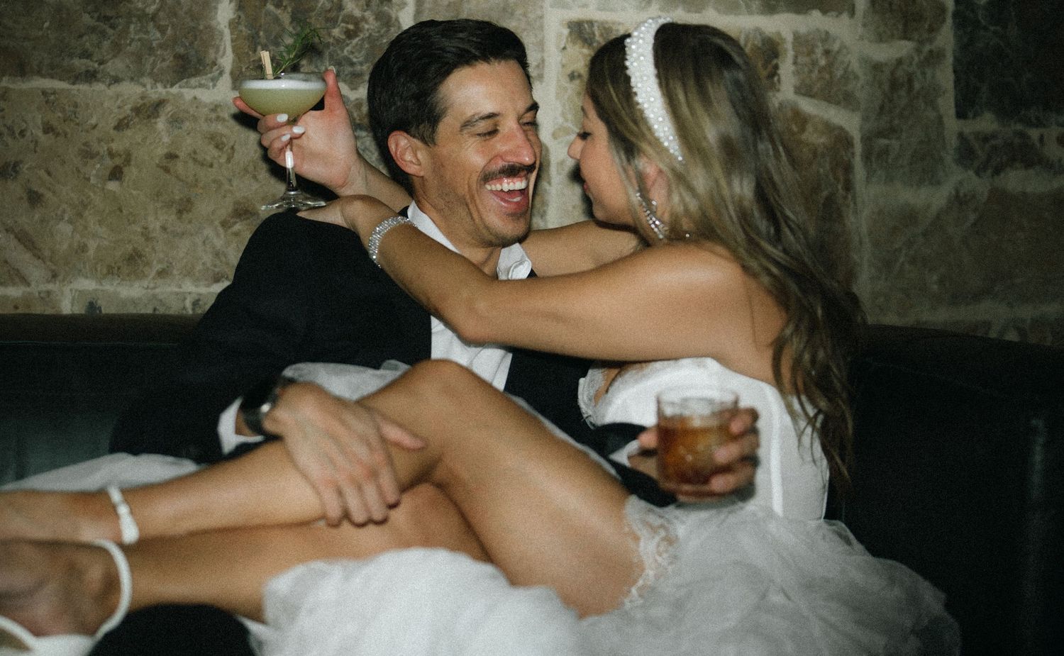 Newlywed couple laughing and cuddling on a couch, holding cocktail glasses in wedding attire.