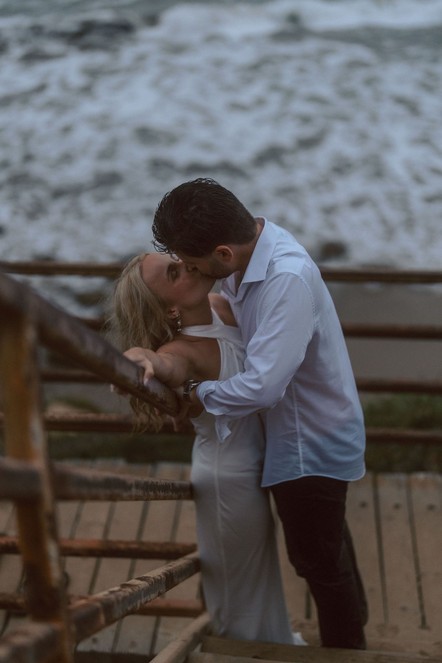 Couple sharing romantic kiss on wooden boardwalk overlooking crashing ocean waves at dusk.