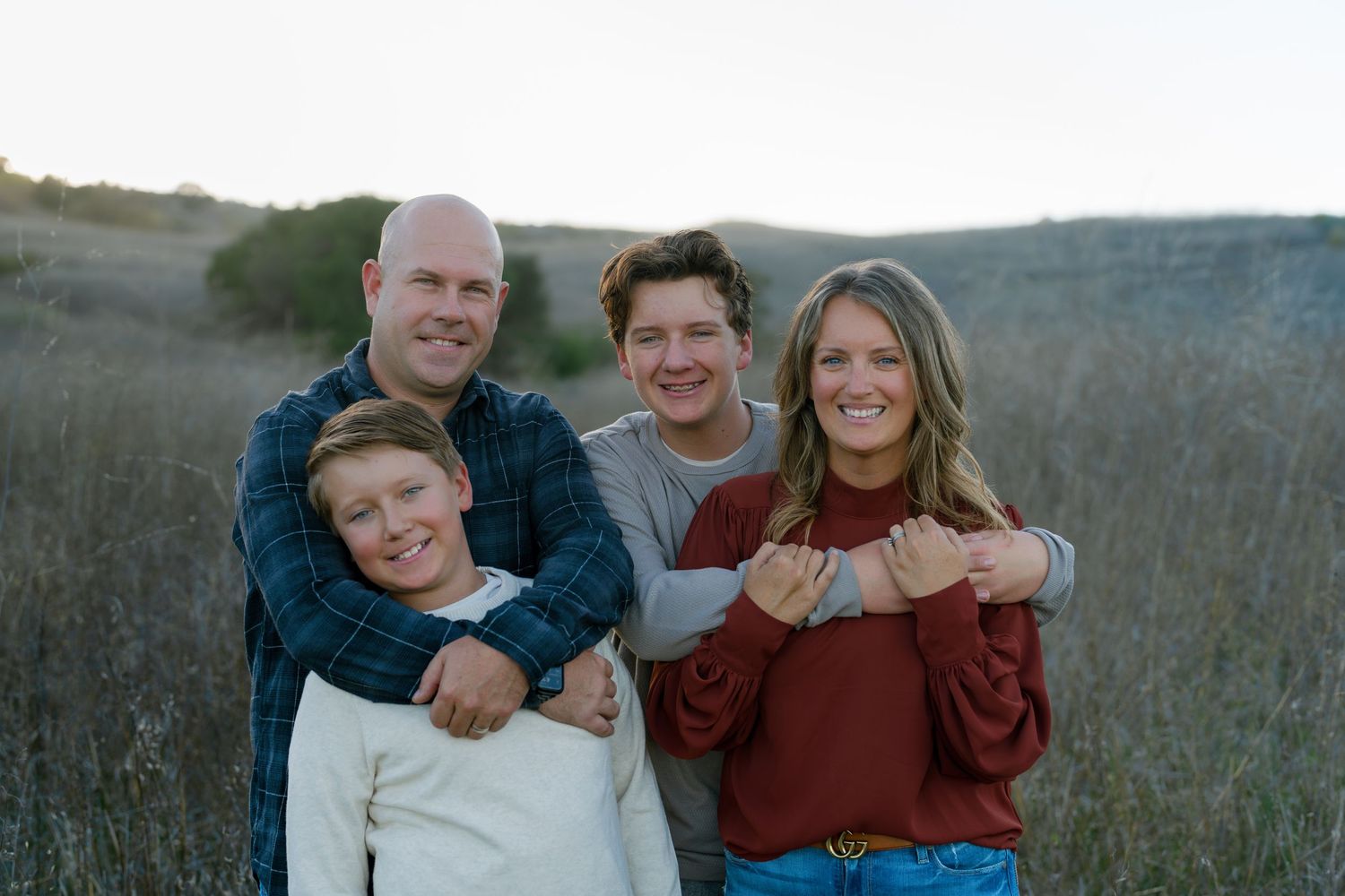 Happy family of four smiling together in a golden field at sunset wearing casual autumn outfits.